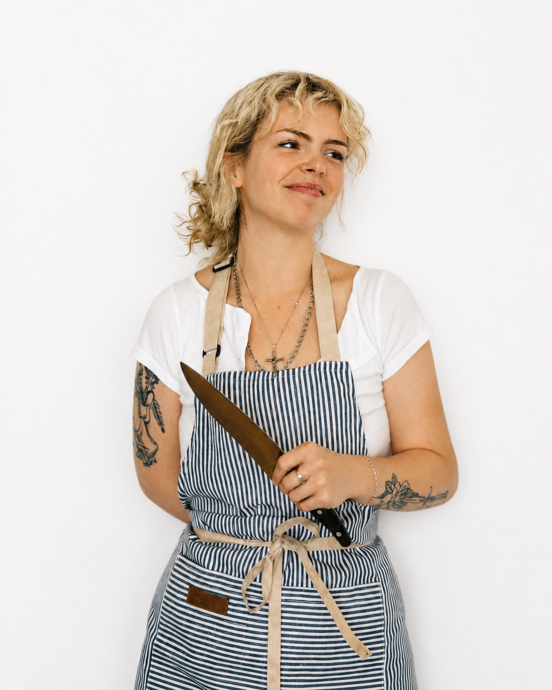 A woman with blonde curly hair holding a chef's knife, wearing an apron with blue and white stripes, a white t-shirt, and jewelry, standing against a plain white background.