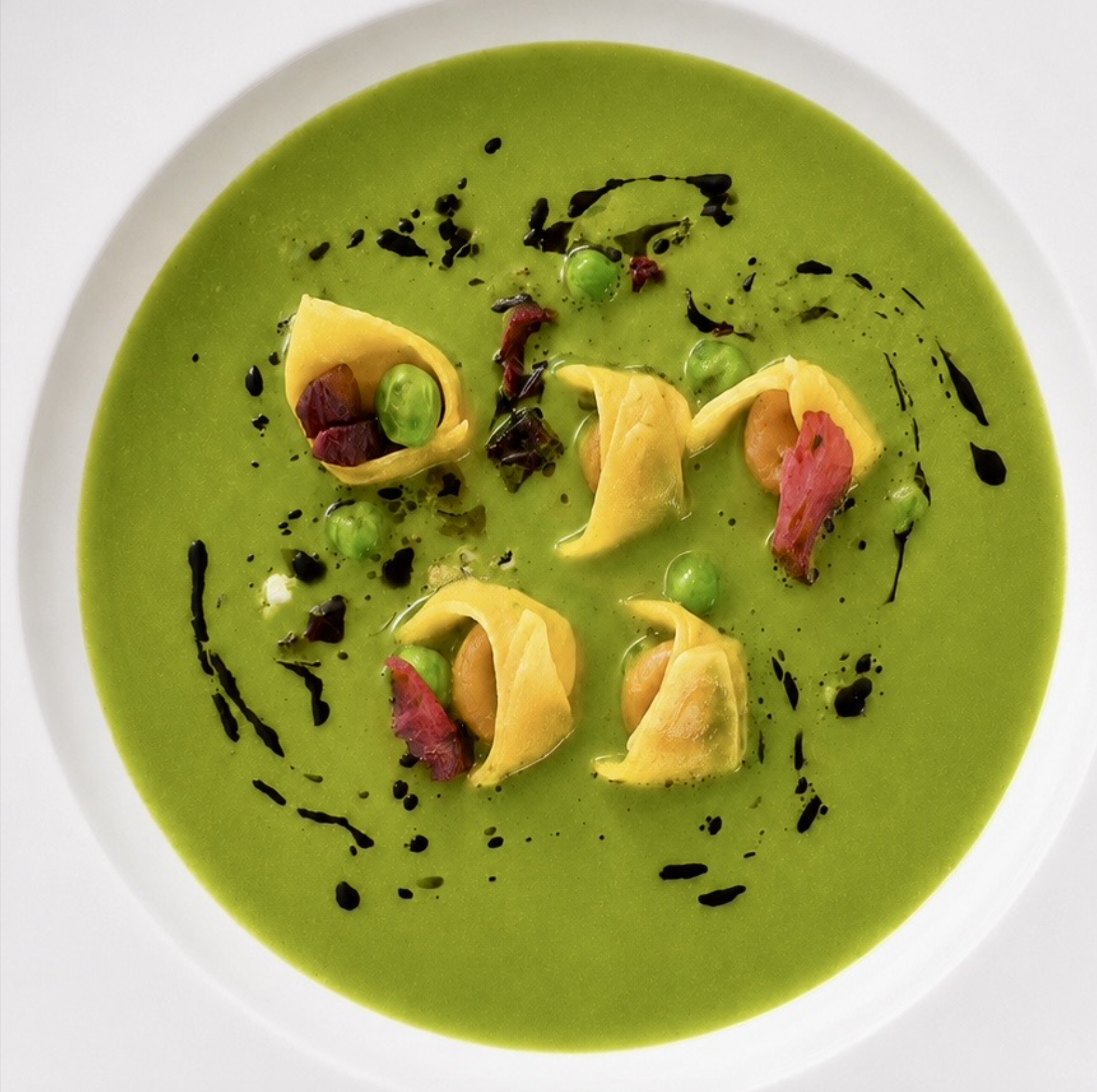 A bowl of bright green soup garnished with yellow pasta, green peas, small pieces of red and brown ingredients, and black drizzle or oil on top.