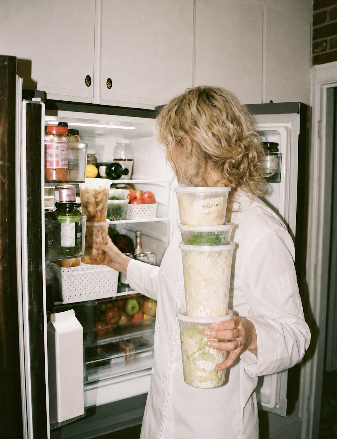 A woman with curly blonde hair wearing a white coat is standing in front of an open refrigerator. She is holding four clear plastic containers filled with chopped vegetables or salads, labeled on the top container. The refrigerator shelf contains various food items, including a bowl of apples, bottles, and jars.
