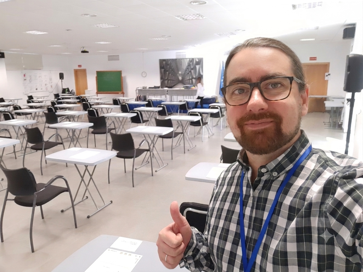 A man with glasses, a beard, and a checkered shirt taking a selfie in an empty conference room, giving a thumbs-up.