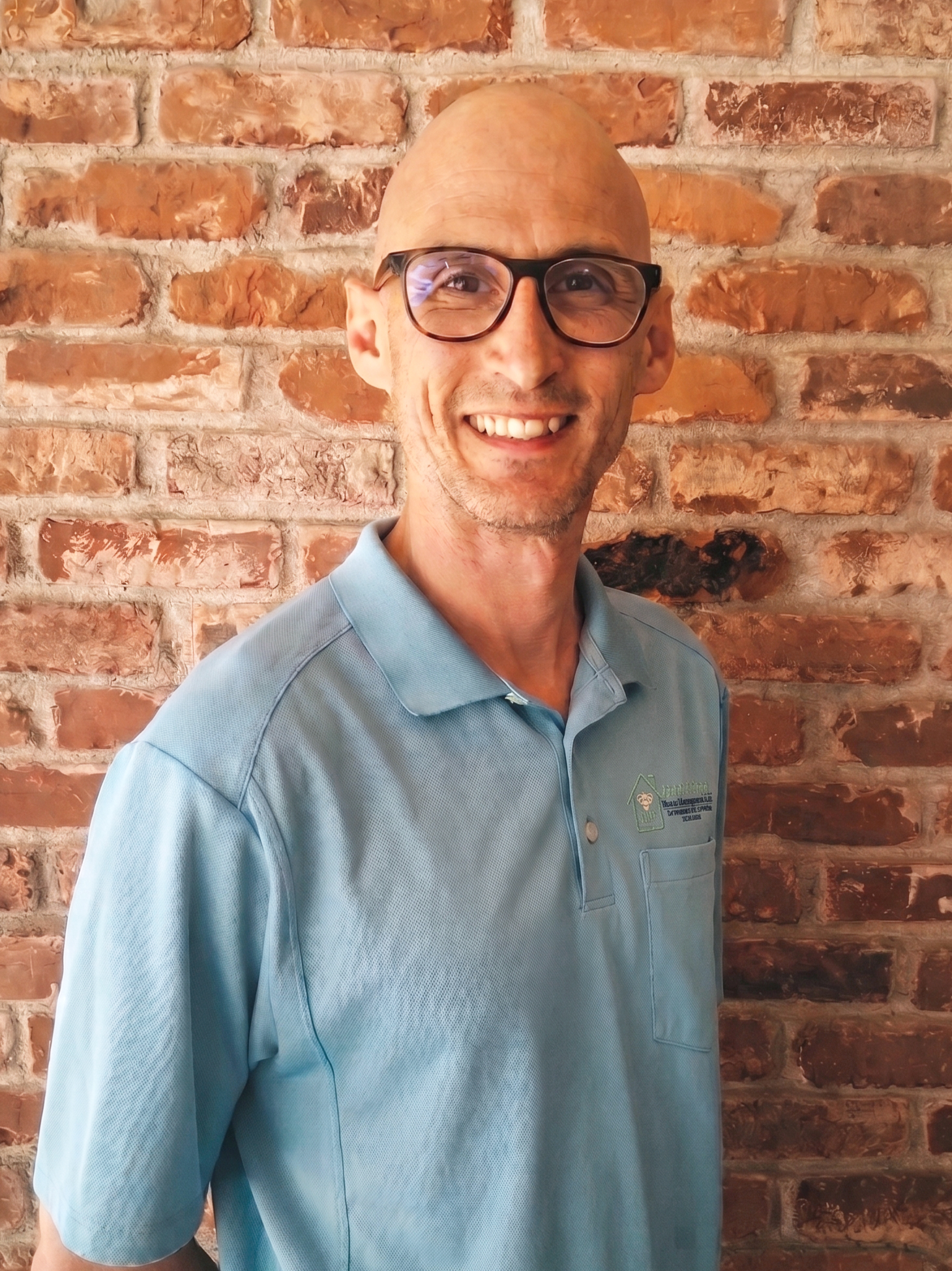 A smiling man wearing glasses and a light blue polo shirt standing in front of a red brick wall.