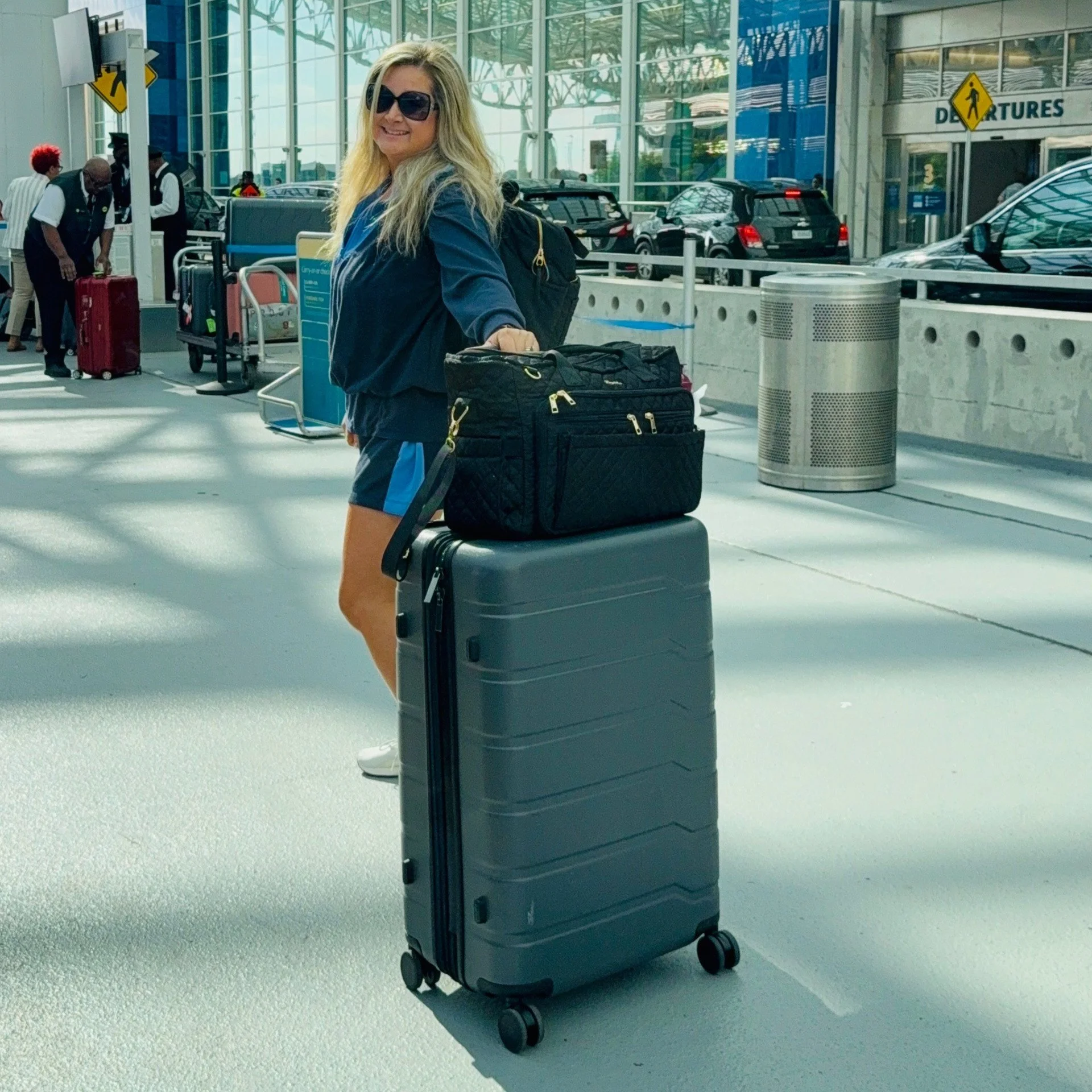 Woman in sunglasses with blonde hair, wearing a dark jacket, smiling, standing at airport baggage claim with rolling suitcase, black carry-on, nearby luggage carts, and people in the background.