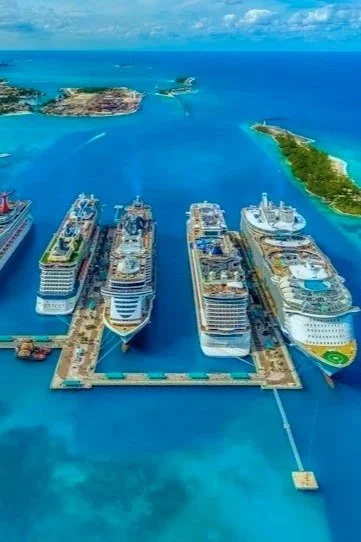 Aerial view of a cruise ship dock with several large cruise ships at a port, surrounded by clear blue water and green islands in the background.