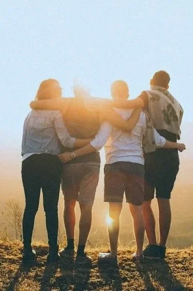 Five friends standing together outdoors at sunset, with their arms around each other's shoulders.