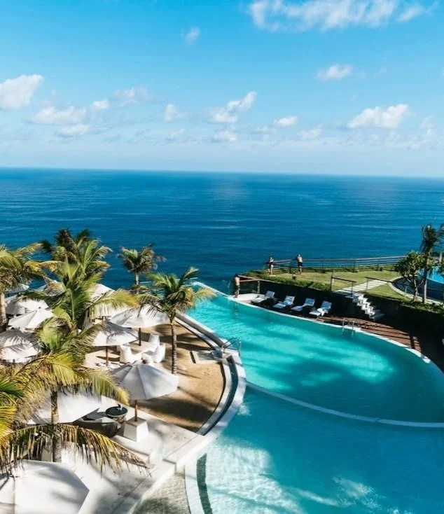 Luxury pool overlooking the ocean with palm trees, lounge chairs, and umbrellas.