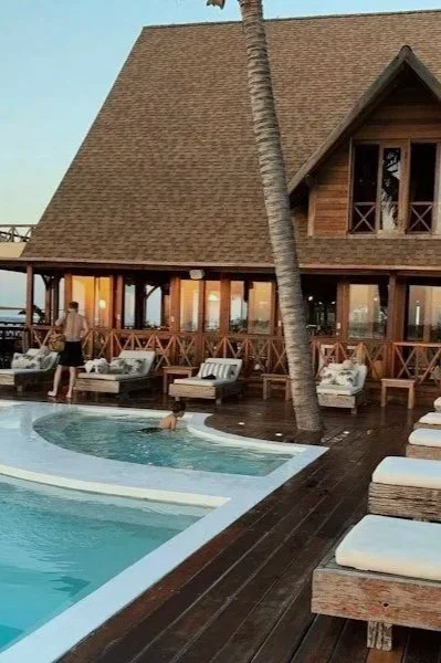 A resort pool area with lounge chairs and a wooden building with large windows and a peaked roof, set near a beach with a palm tree.