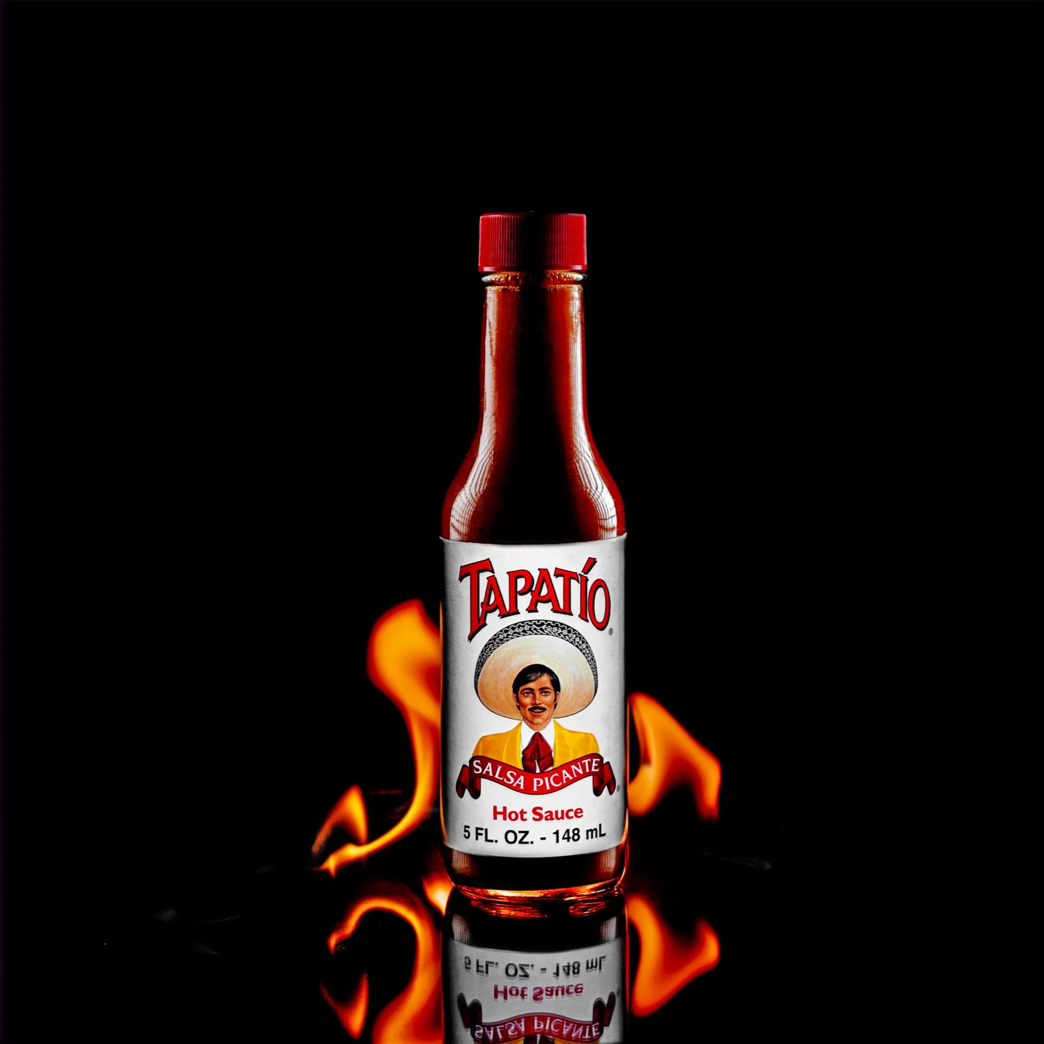 A bottle of Tapatio hot sauce with flames and a reflection of the label visible. The label features a man in a yellow suit with a red tie, and the brand name 'Tapatio' in red letters.