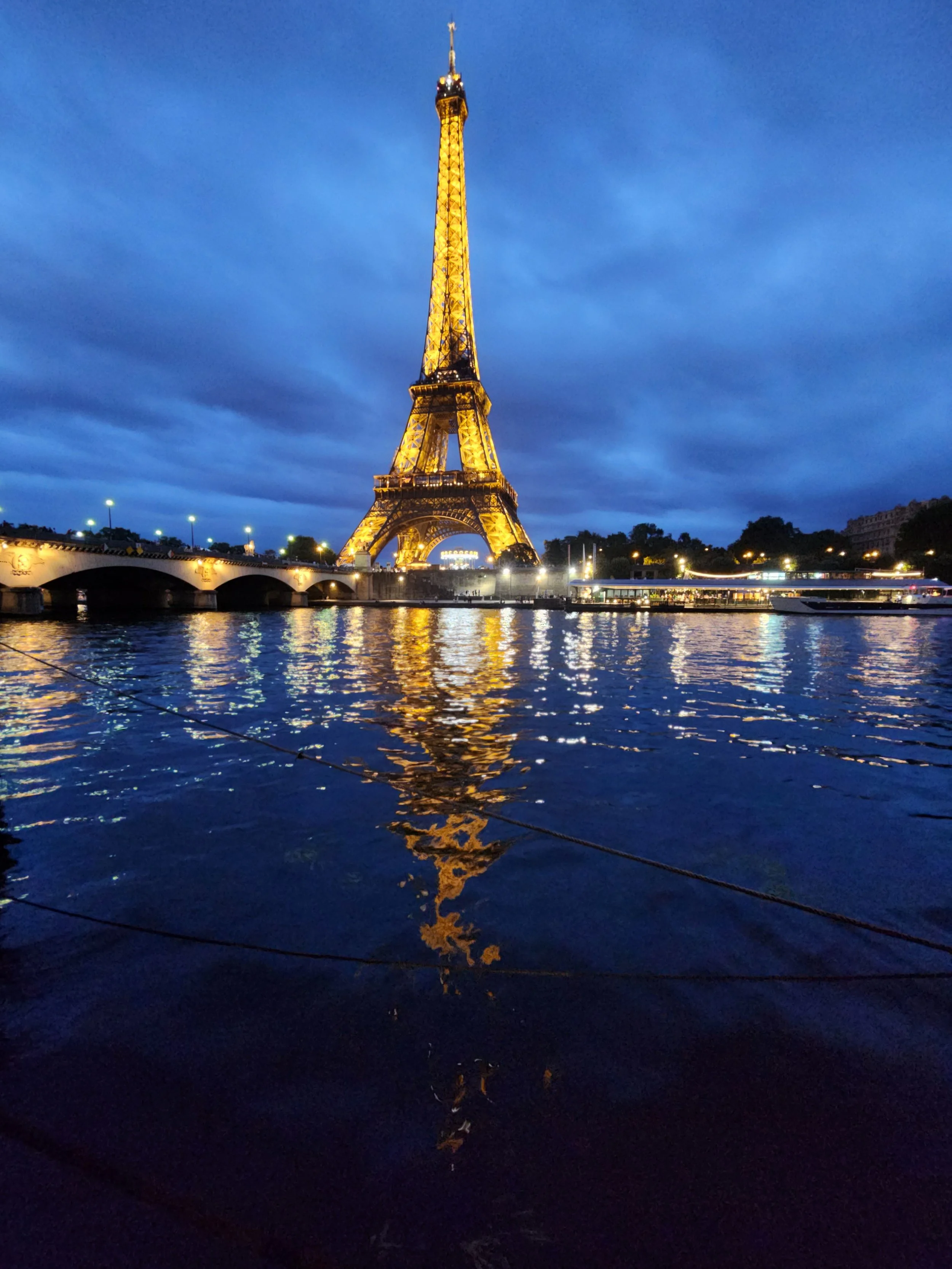 The Eiffel Tower illuminated with yellow lights at dusk, with a reflective water body in the foreground and a cloudy blue sky overhead.