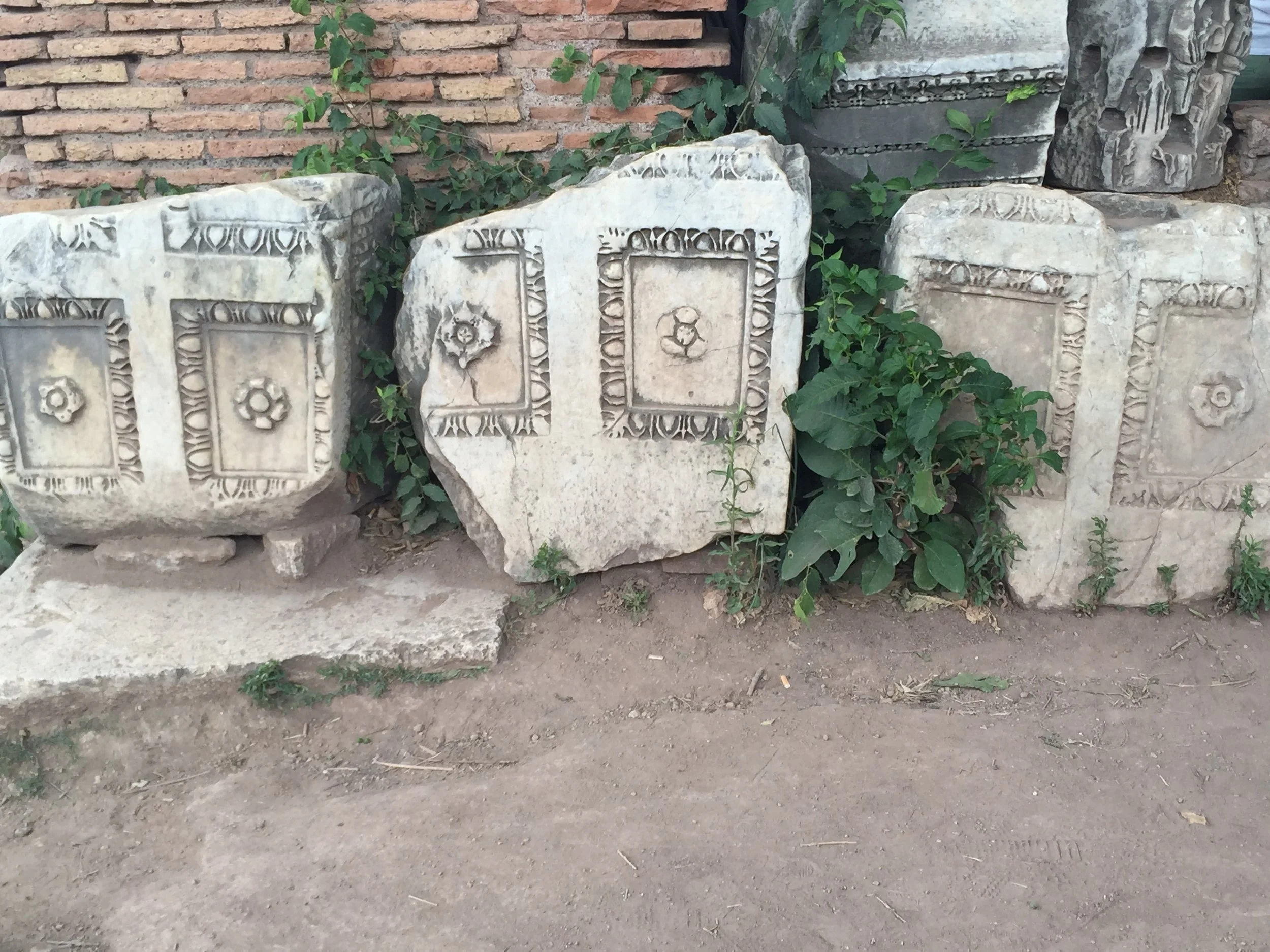 Ancient stone blocks with carved decorative patterns stacked against a brick wall, some overgrown with green ivy.