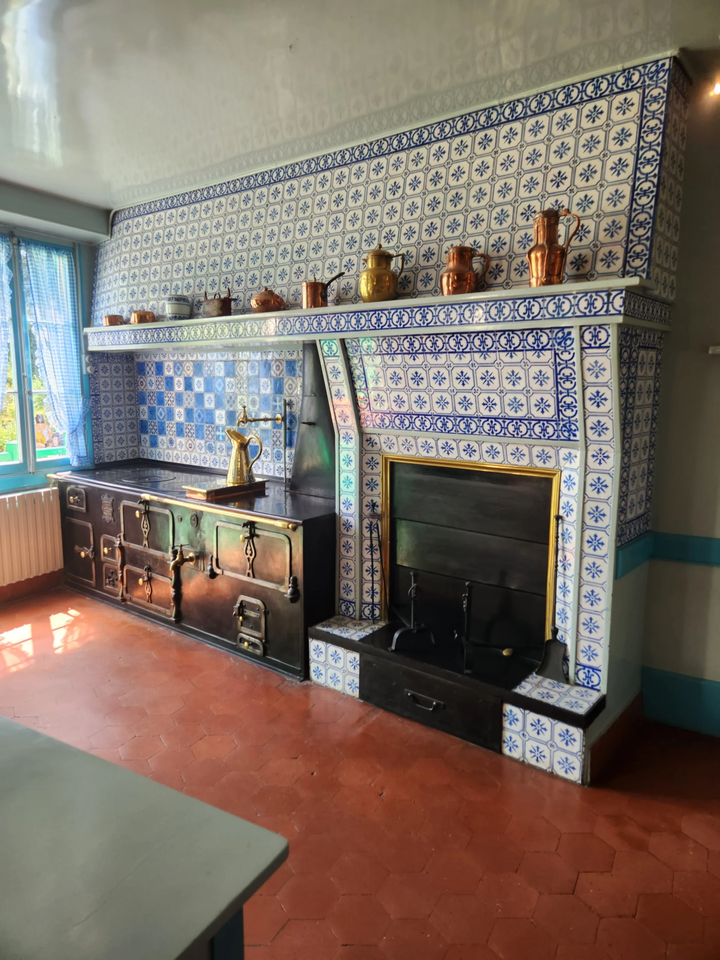 A traditional kitchen with a vintage black stove, brass faucet, and a white tiled wall with blue decorative patterns. There is a window with blue gingham curtains on the left and various copper and brass pots on a shelf above the stove.