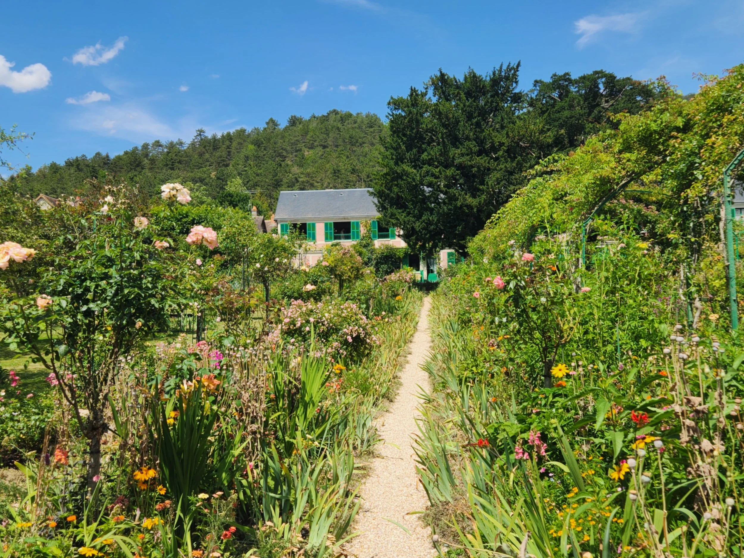 A garden with pink, orange, and yellow flowers along a gravel path leading to a house with green window shutters. Tall trees and a hillside in the background under a partly cloudy blue sky.