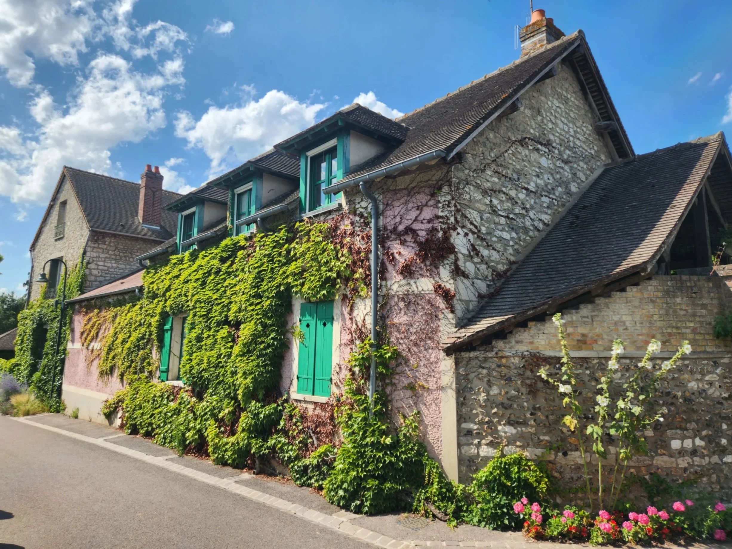 A row of houses with stone walls and various roof styles, some with green shutters, greenery, and pink flowers along the sidewalk, under a partly cloudy blue sky.