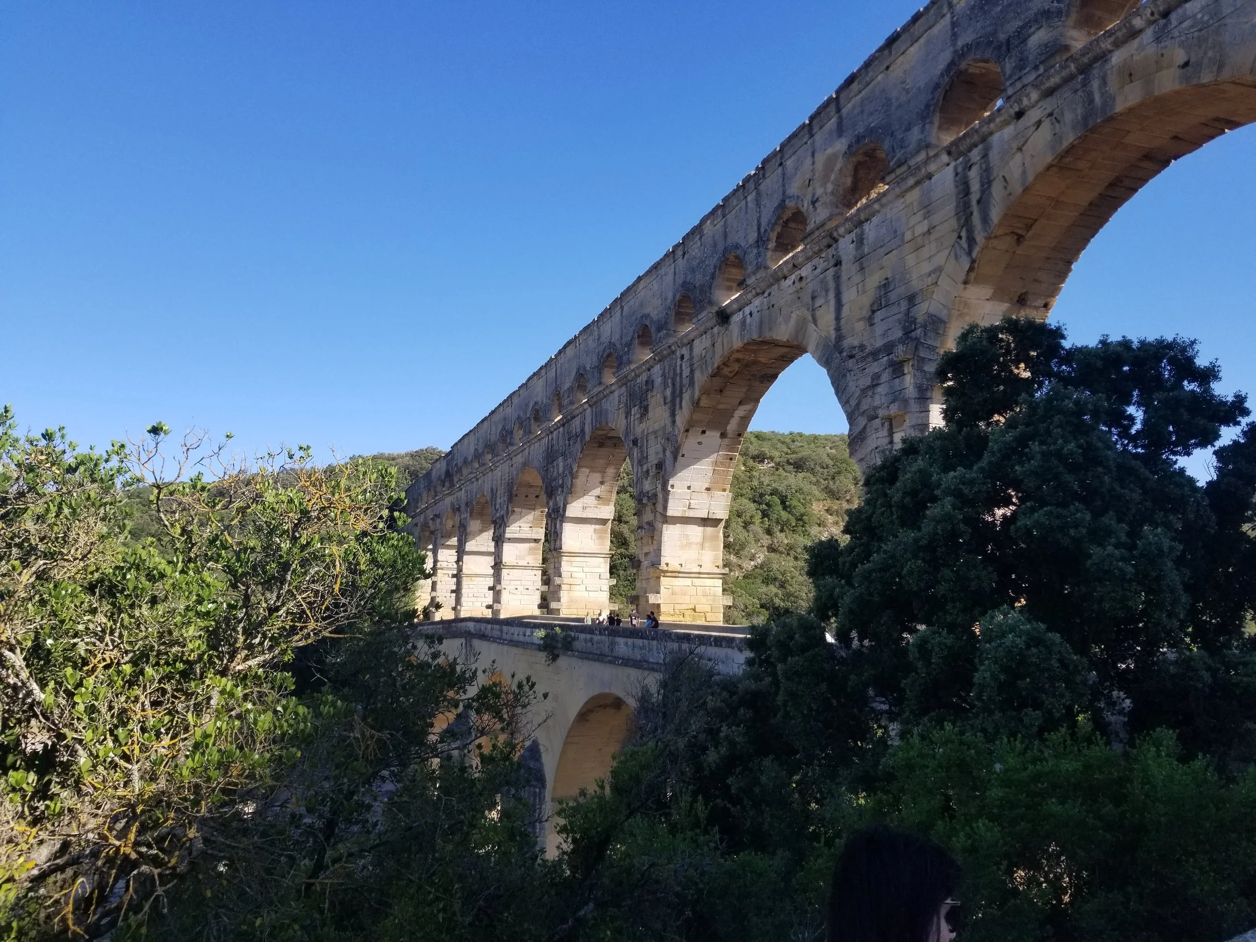 An ancient Roman aqueduct with multiple arches, surrounded by greenery and trees, under a clear blue sky.