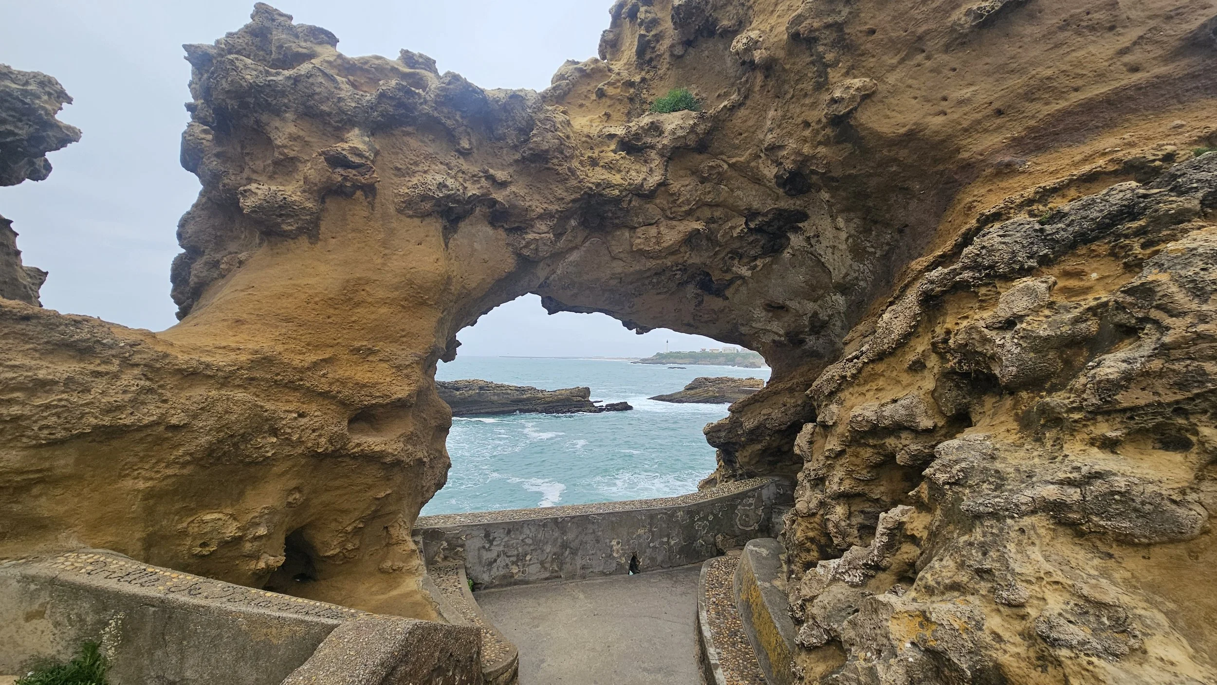Natural rock formation with a hole in the middle, overlooking the ocean with small waves and a distant shoreline, sky is partly cloudy.