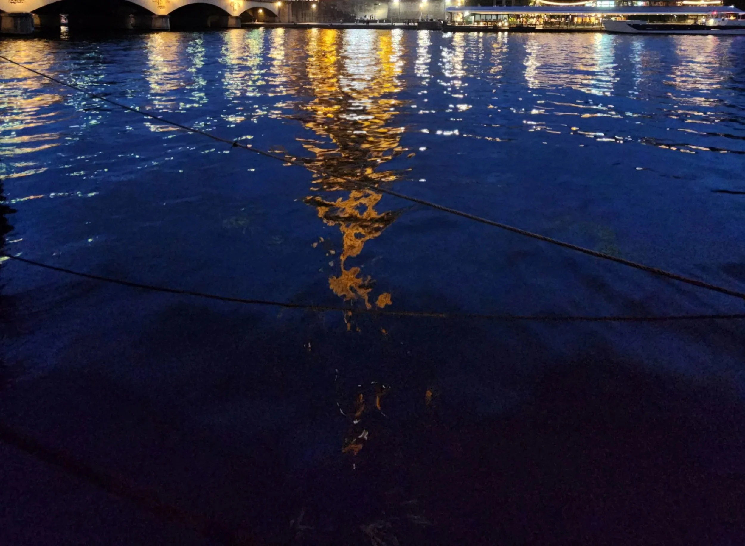 Nighttime scene of a body of water reflecting colorful lights from the cityscape and a bridge in the background, with boats docked nearby.