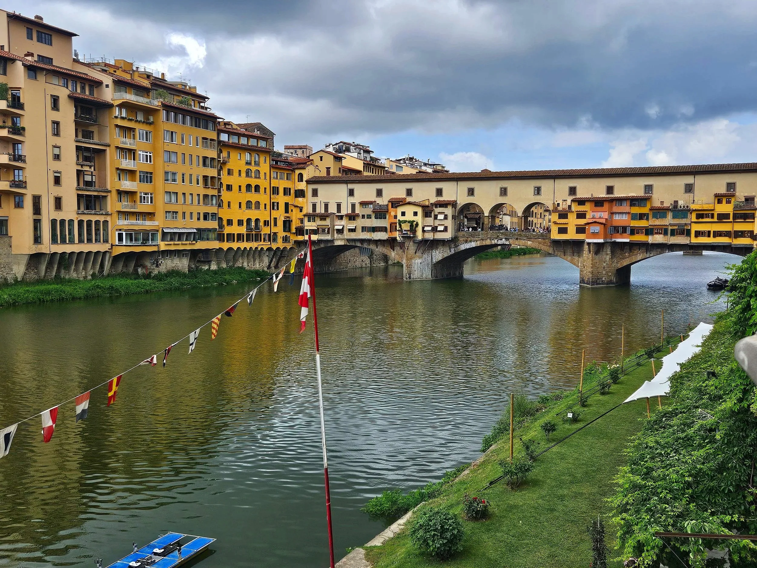 Colorful buildings along a river with a bridge and boats, dark clouds in the sky, flags hanging across the river, lush green grass and plants on the riverbank.