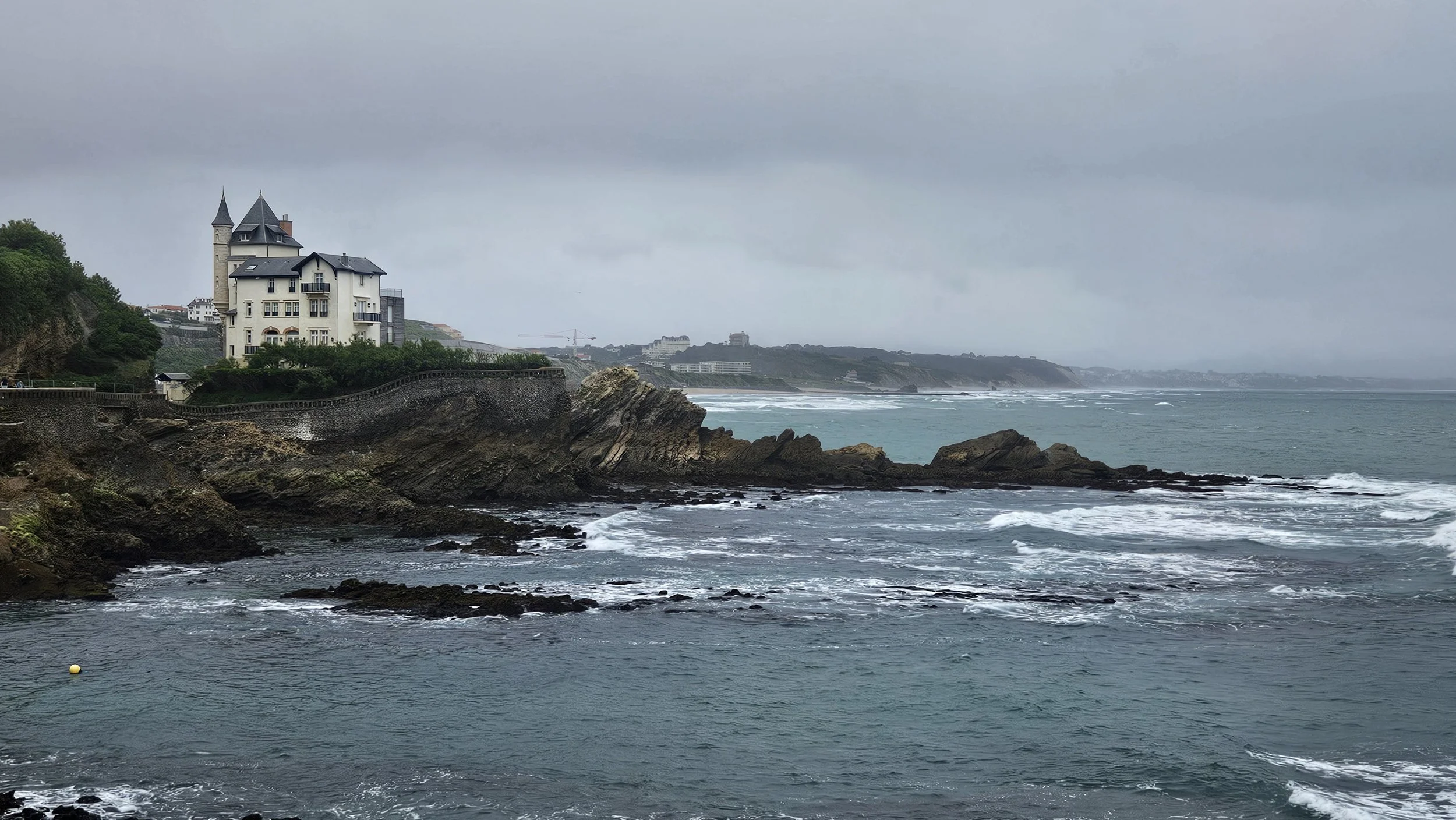 Coastal landscape featuring a large white building with towers on a cliff over the ocean under a cloudy sky.