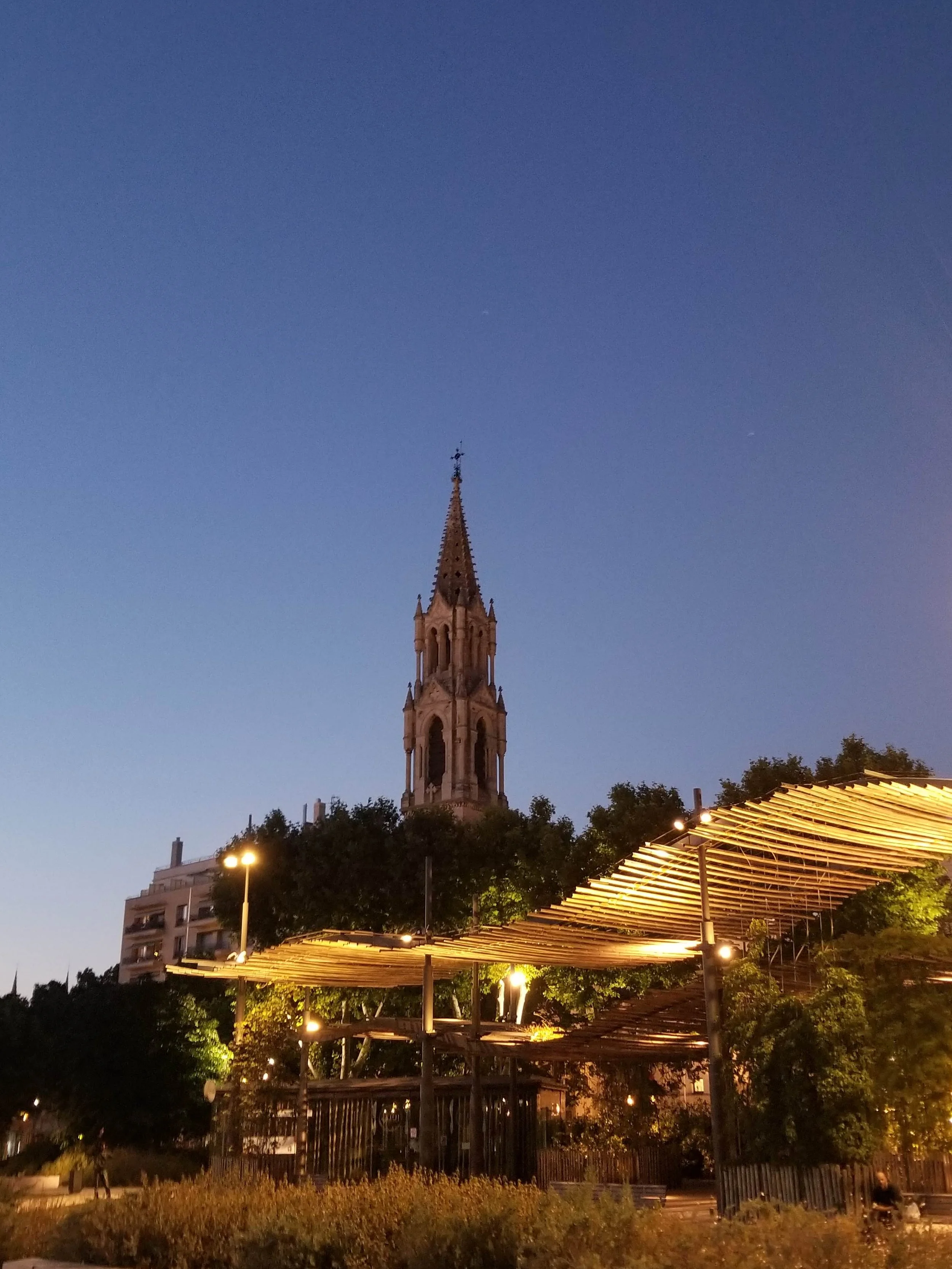 Nimes at Night - A tall church steeple with clock faces, surrounded by trees and illuminated structures, under a clear evening sky