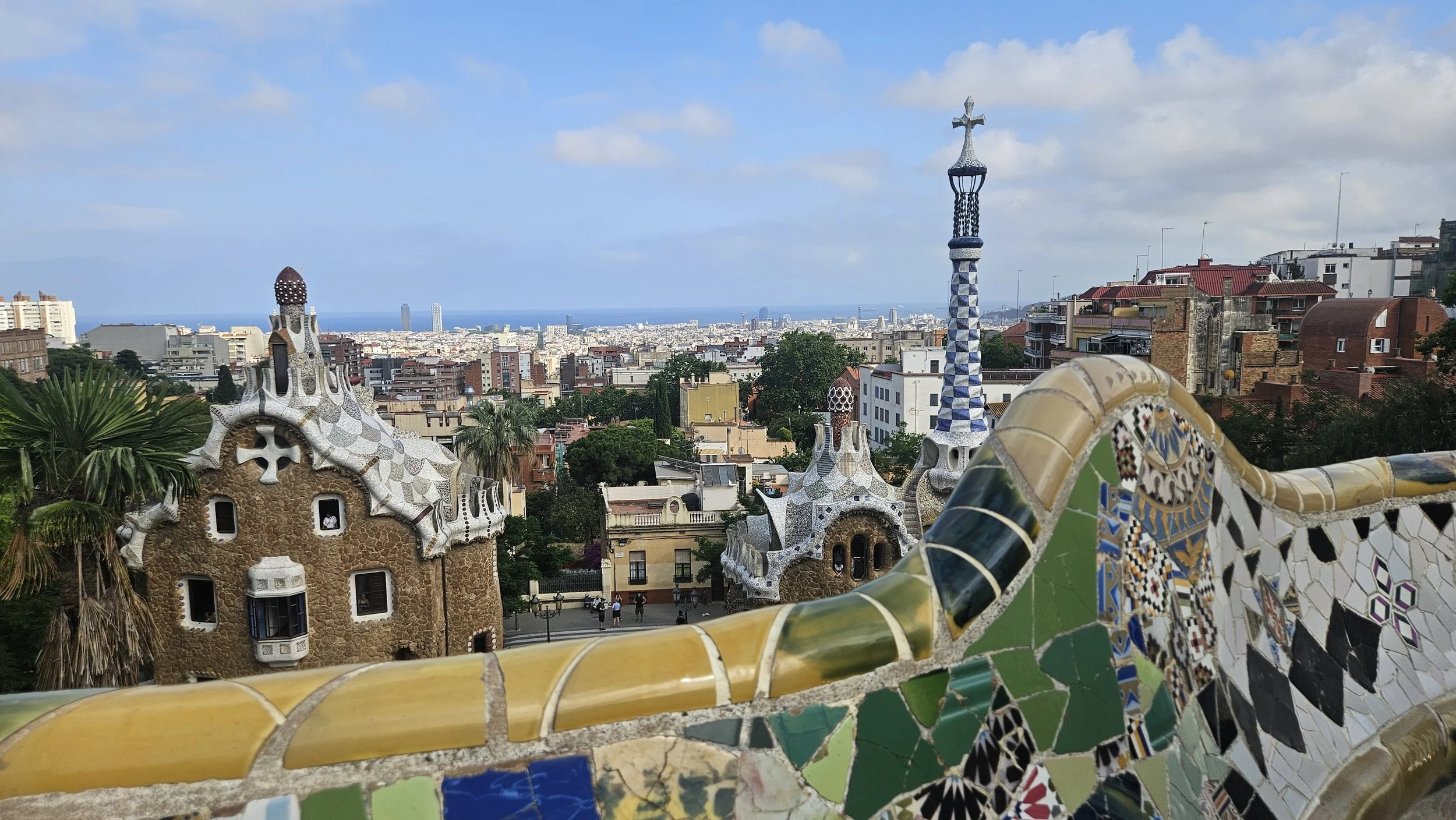 Colorful ceramic mosaic railing overlooking Park Güell in Barcelona, with whimsical mosaic buildings featuring curvy shapes and tile details, cityscape in the background, and a partly cloudy sky.