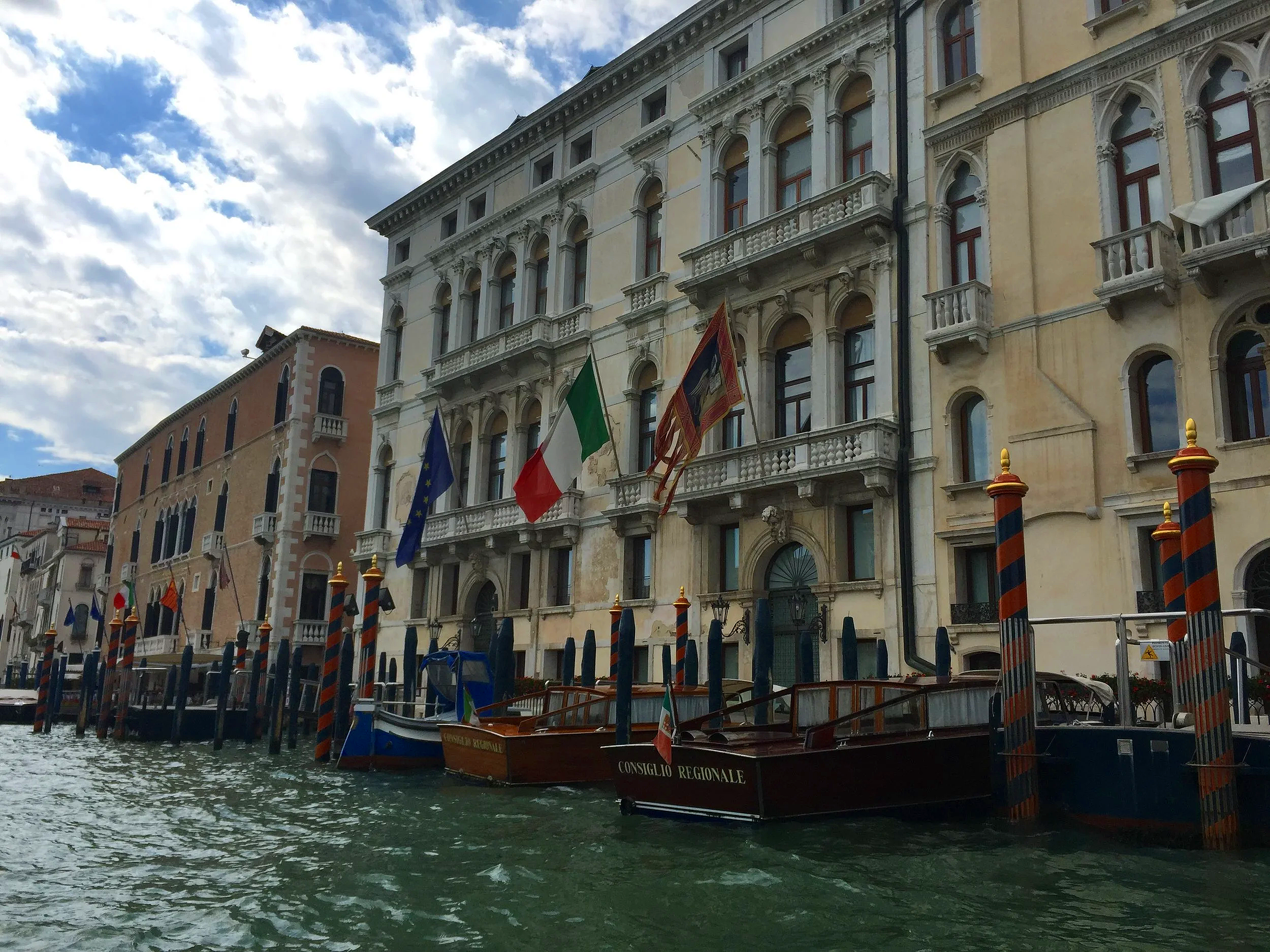 Buildings along a canal, with boats docked in the water and flags hanging from the buildings. The sky is partly cloudy.