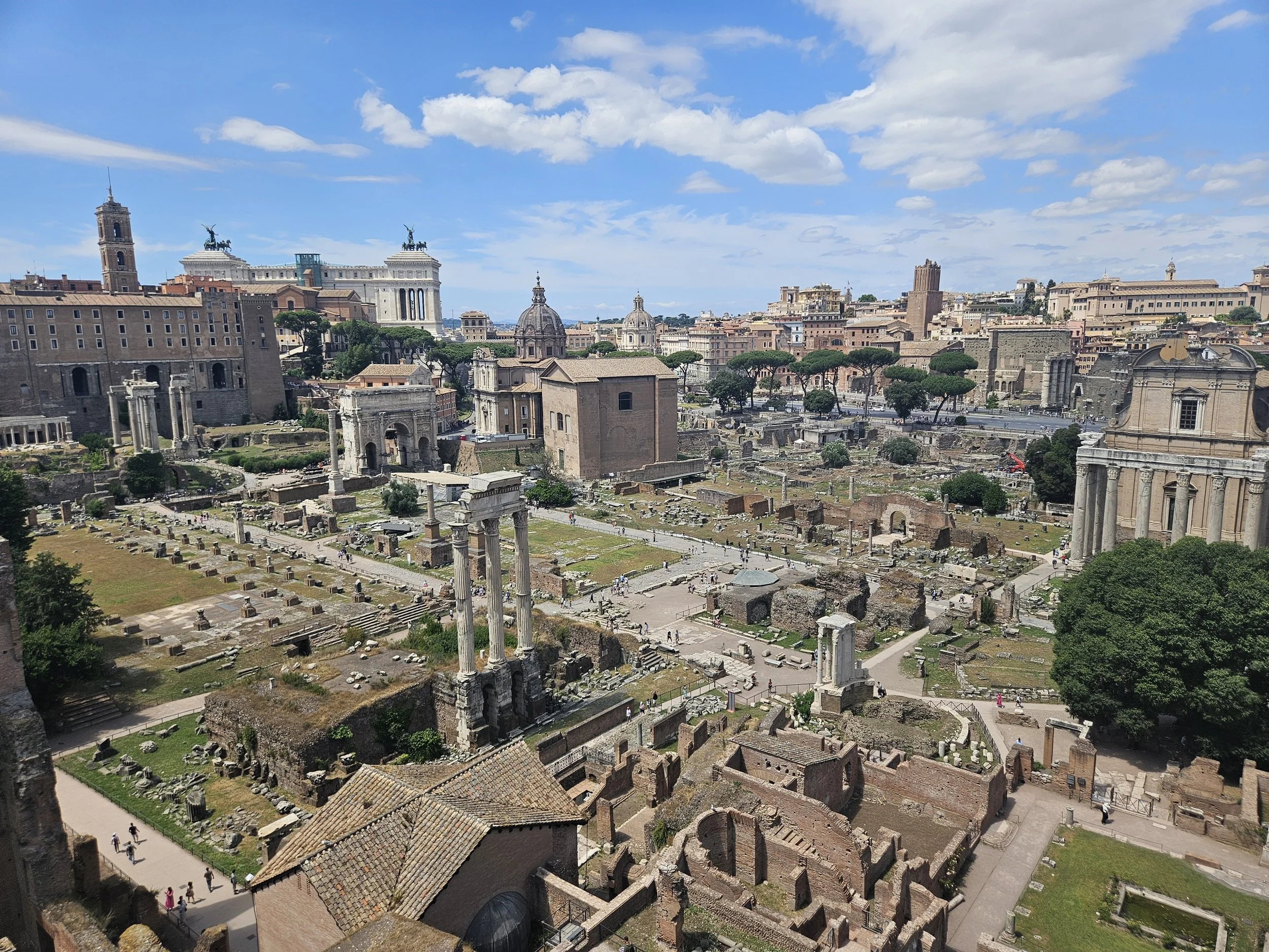 A panoramic view of ancient ruins and historical buildings in Rome, Italy, with a blue sky and scattered clouds.