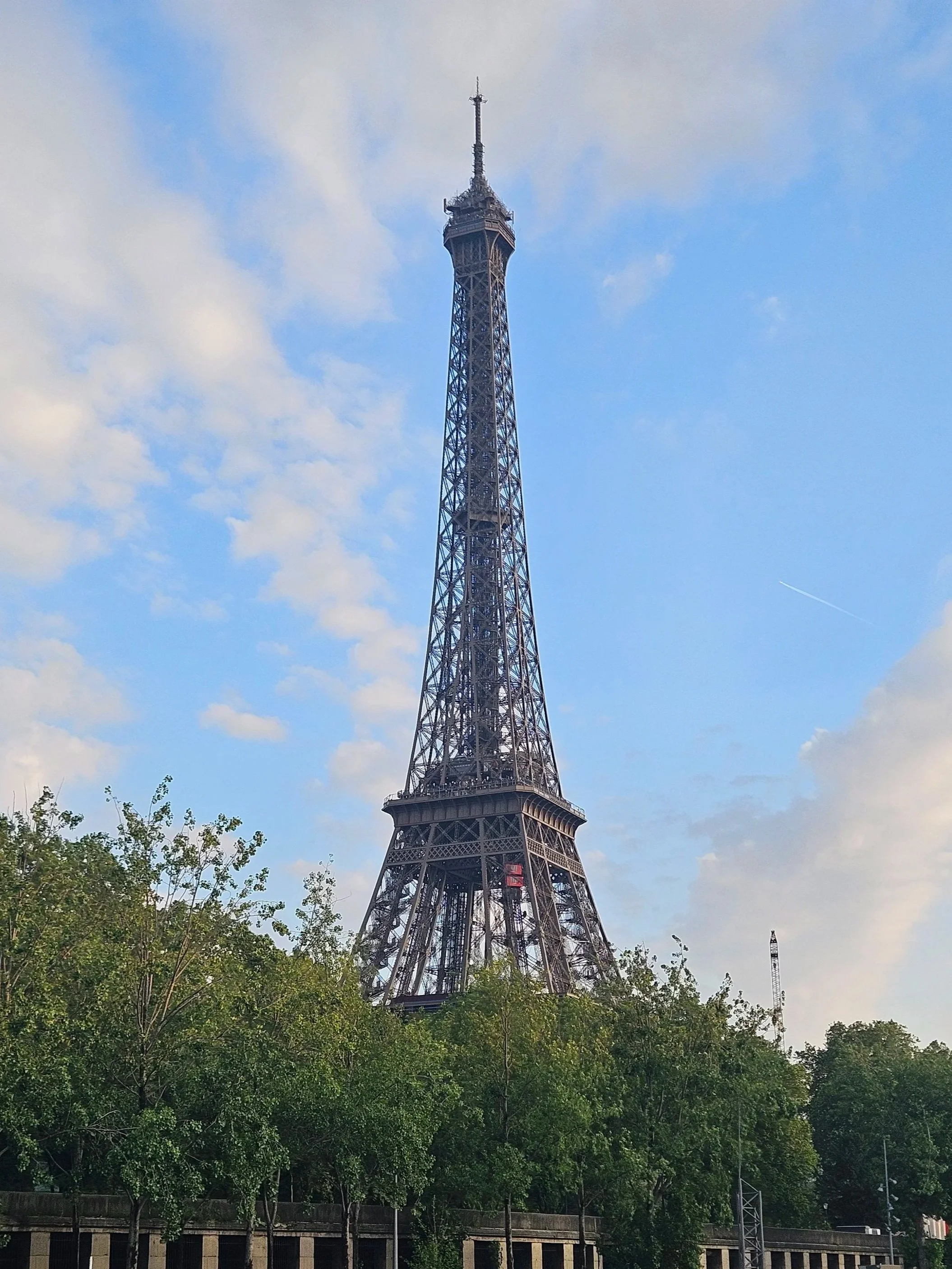 Eiffel Tower from the Seine - the Eiffel Tower, seen from a distance with a blue sky and some clouds in the background and trees at the bottom front.