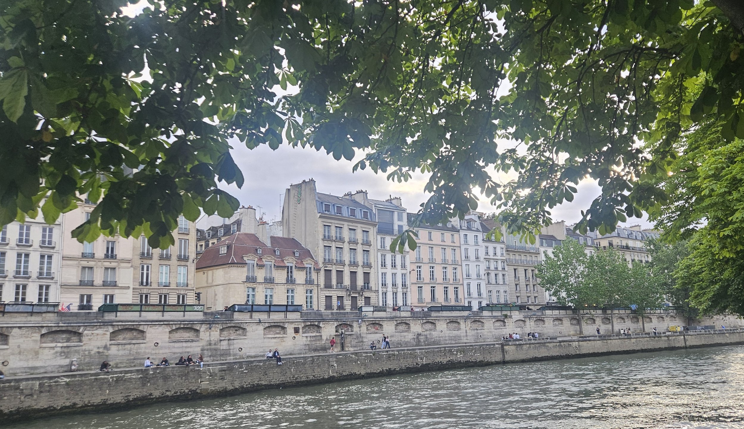 View of European-style buildings along a waterfront, with trees and green foliage in the foreground and a cloudy sky overhead.