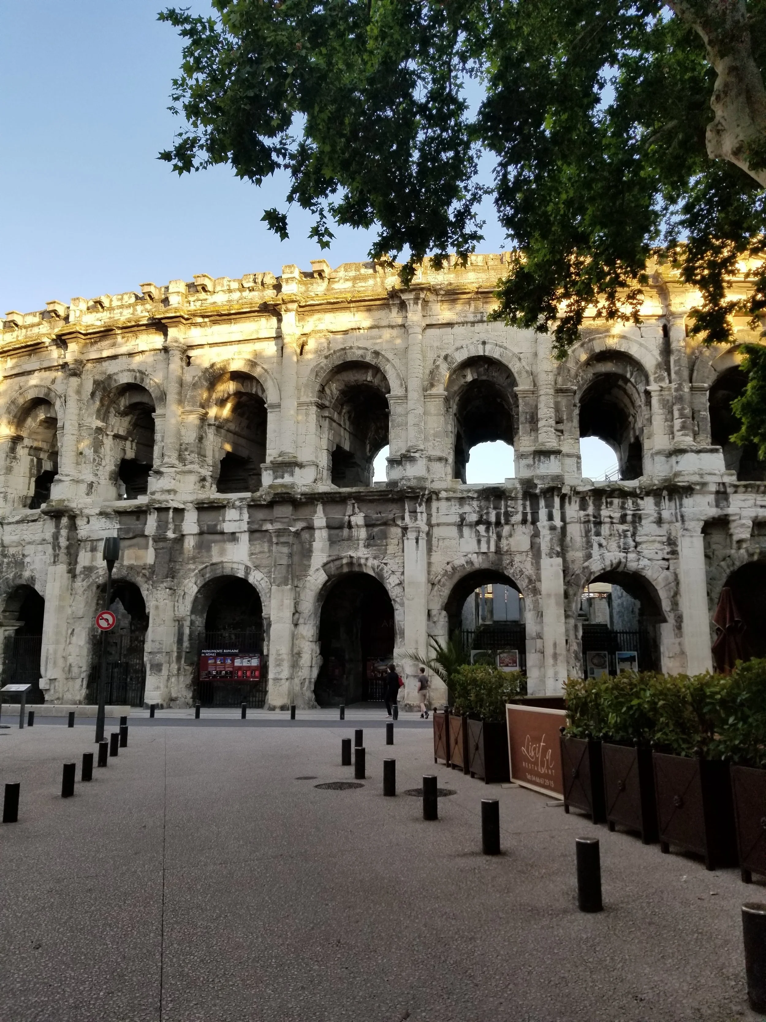 Nimes, France - one of many Roman coliseums built during their rule, seen during daylight with trees overhead and a few people walking nearby.
