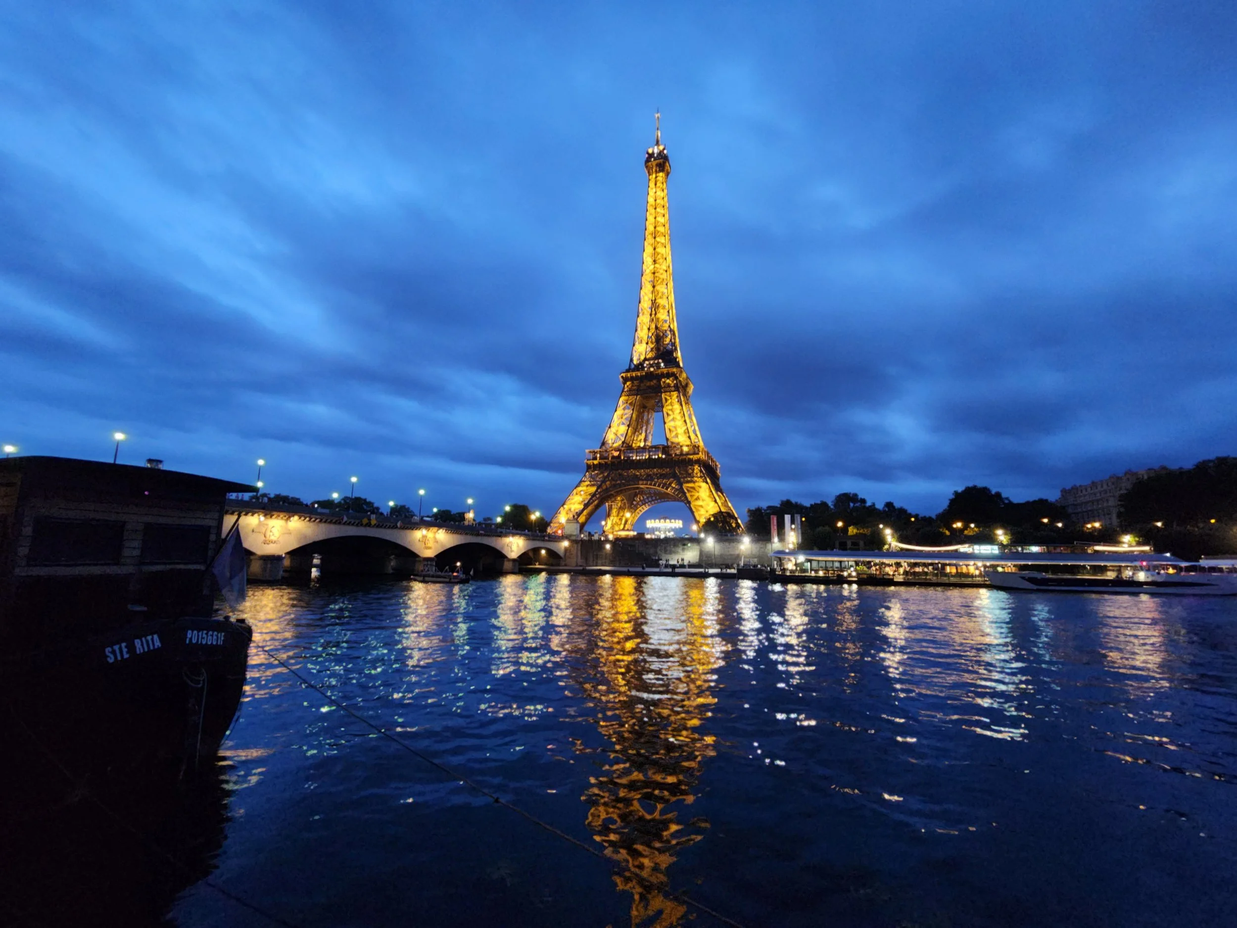The Eiffel Tower illuminated at night with reflections on the water below, set against a dark blue cloudy sky.