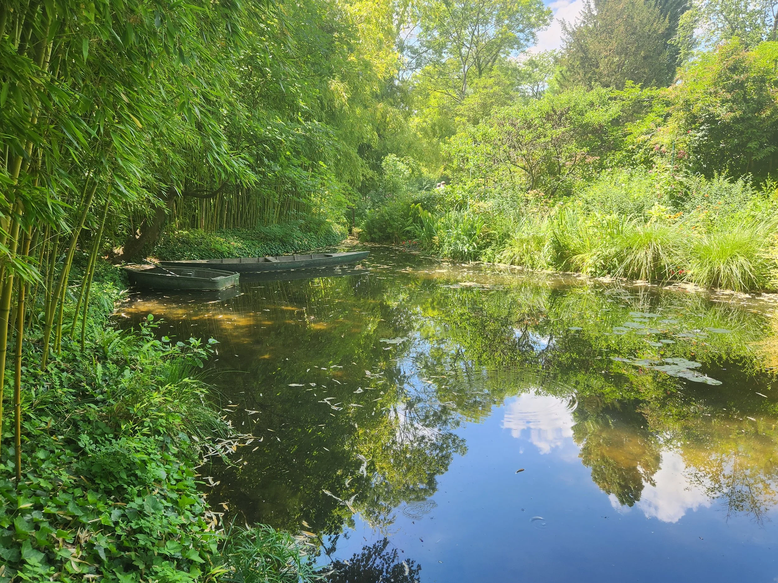 A lush green garden with trees and bushes surrounding a small peaceful pond reflecting the sky and foliage. Two boats are floating on the water.