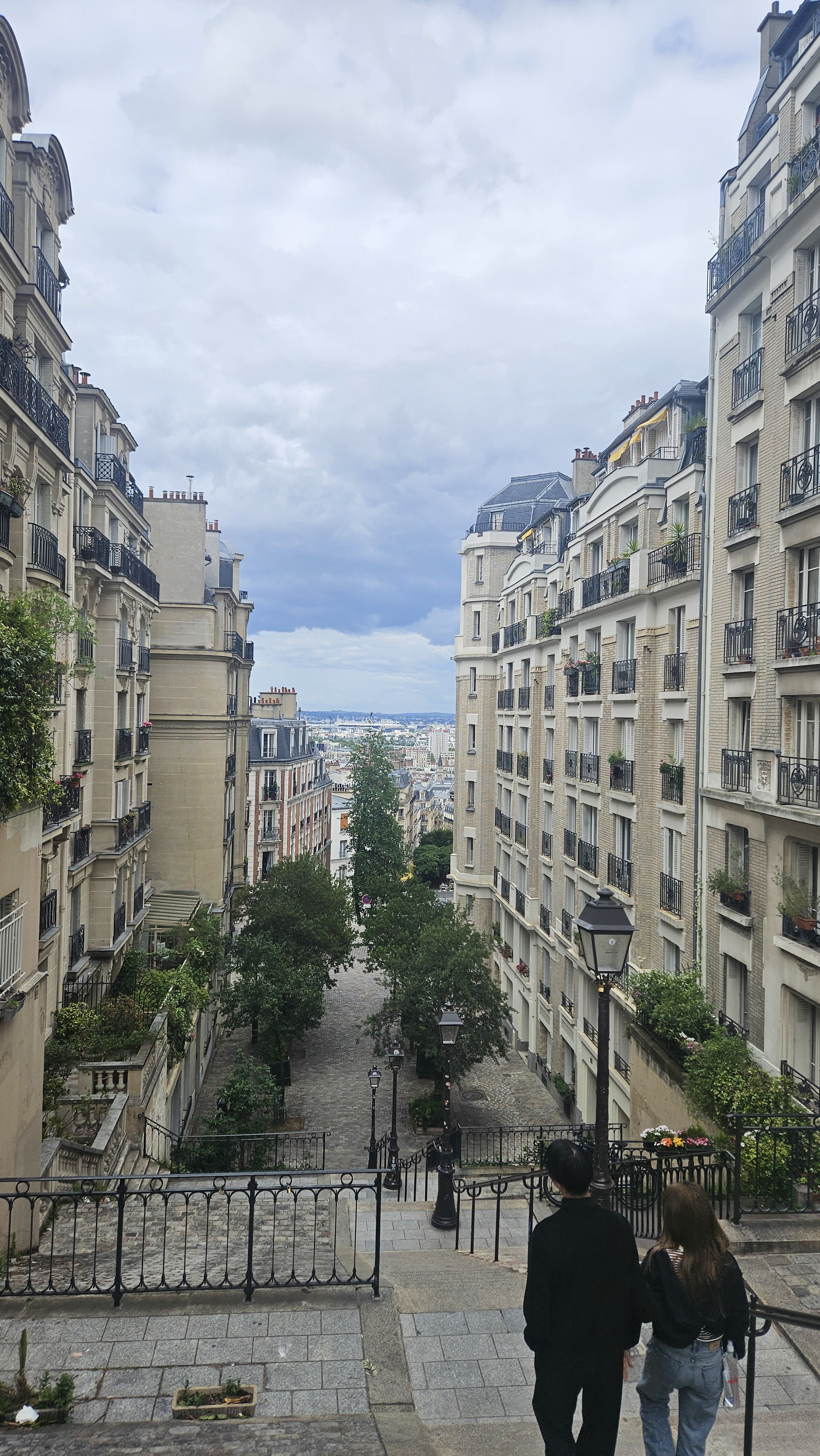 View of a city street flanked by classic European-style apartment buildings with balconies, looking down a cobblestone walkway lined with trees and lampposts, under a cloudy sky, with two people walking down the stairs in the foreground.