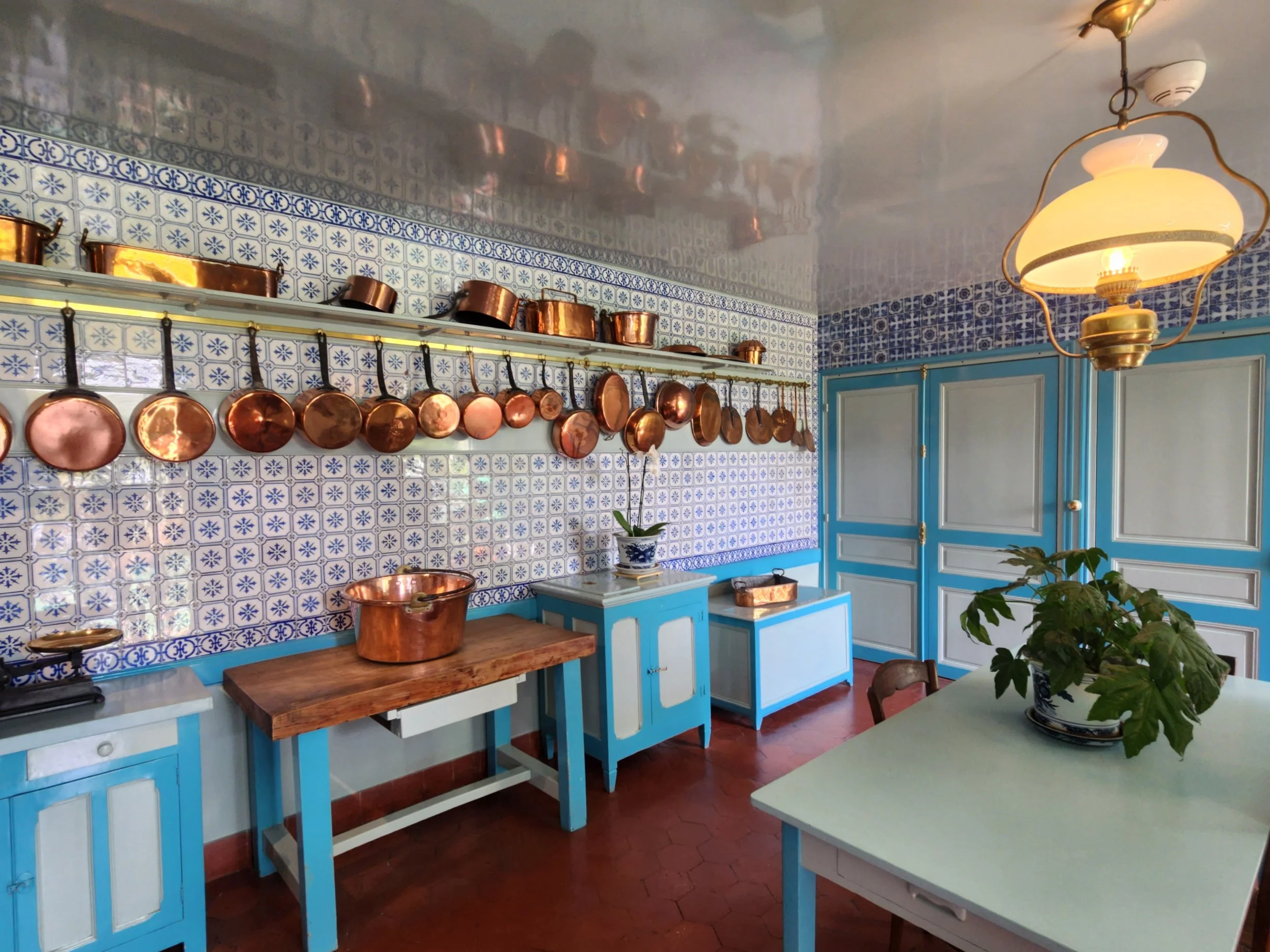 A kitchen with blue cabinets, copper pots hanging on a rail, and patterned white and blue tiled walls. There are wooden and metal countertops, a potted plant, and a vintage ceiling light fixture.