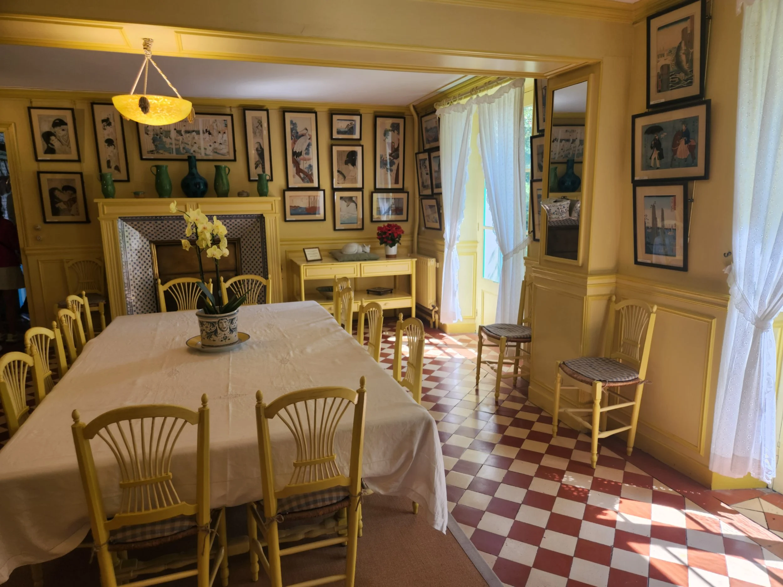 A dining room with a long table covered with a beige tablecloth, surrounded by yellow wooden chairs with plaid cushions. A potted orchid is on the table. The back wall features numerous framed artwork and vases displayed on a yellow mantel. There are sheer white curtains hanging by a window or door, allowing natural light to brighten the room. The floor has a red and beige checkerboard tile pattern.