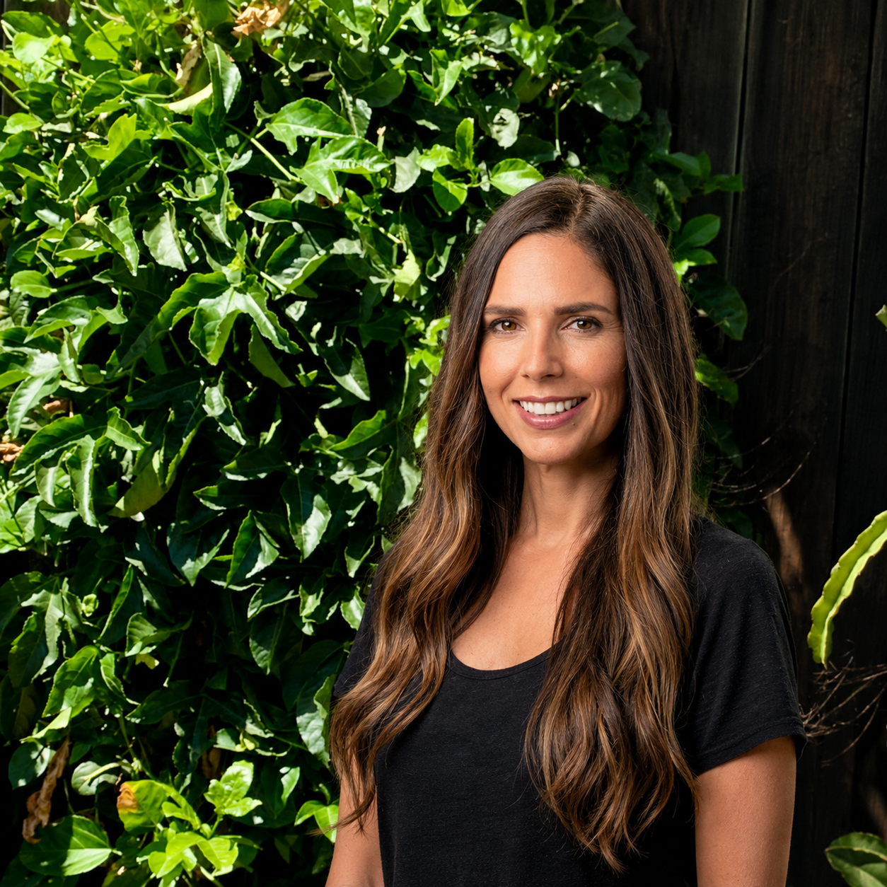 A woman with long wavy brown hair smiling in front of lush green foliage and a dark wooden fence.