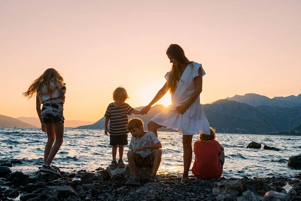 A woman with four children on a rocky beach at sunset, holding hands and playing near the water with mountains in the background.