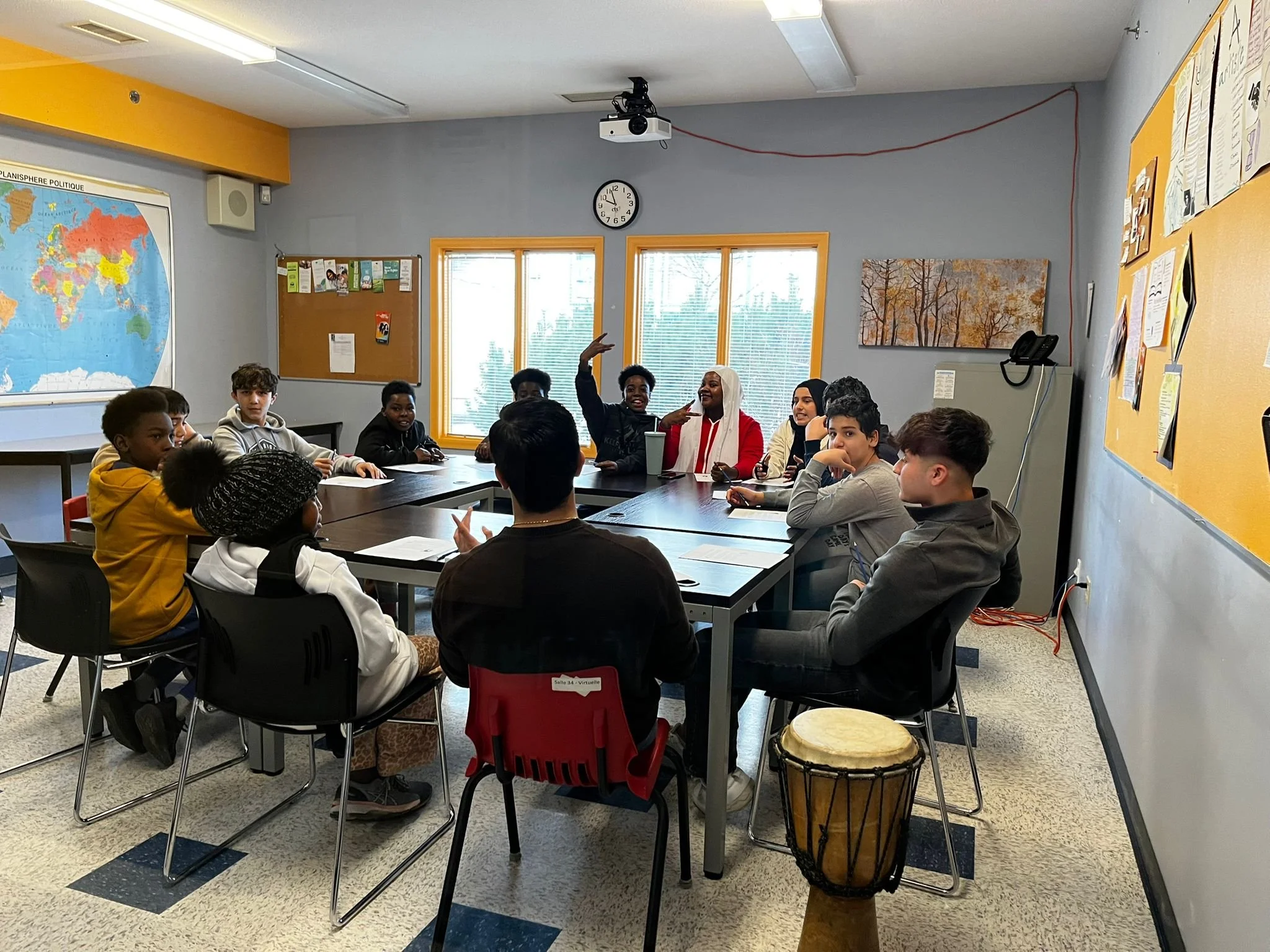 A classroom with students sitting around a table, some engaging in conversation, others looking at the camera, with a world map and bulletin board on the walls, and a clock and projector on the ceiling.