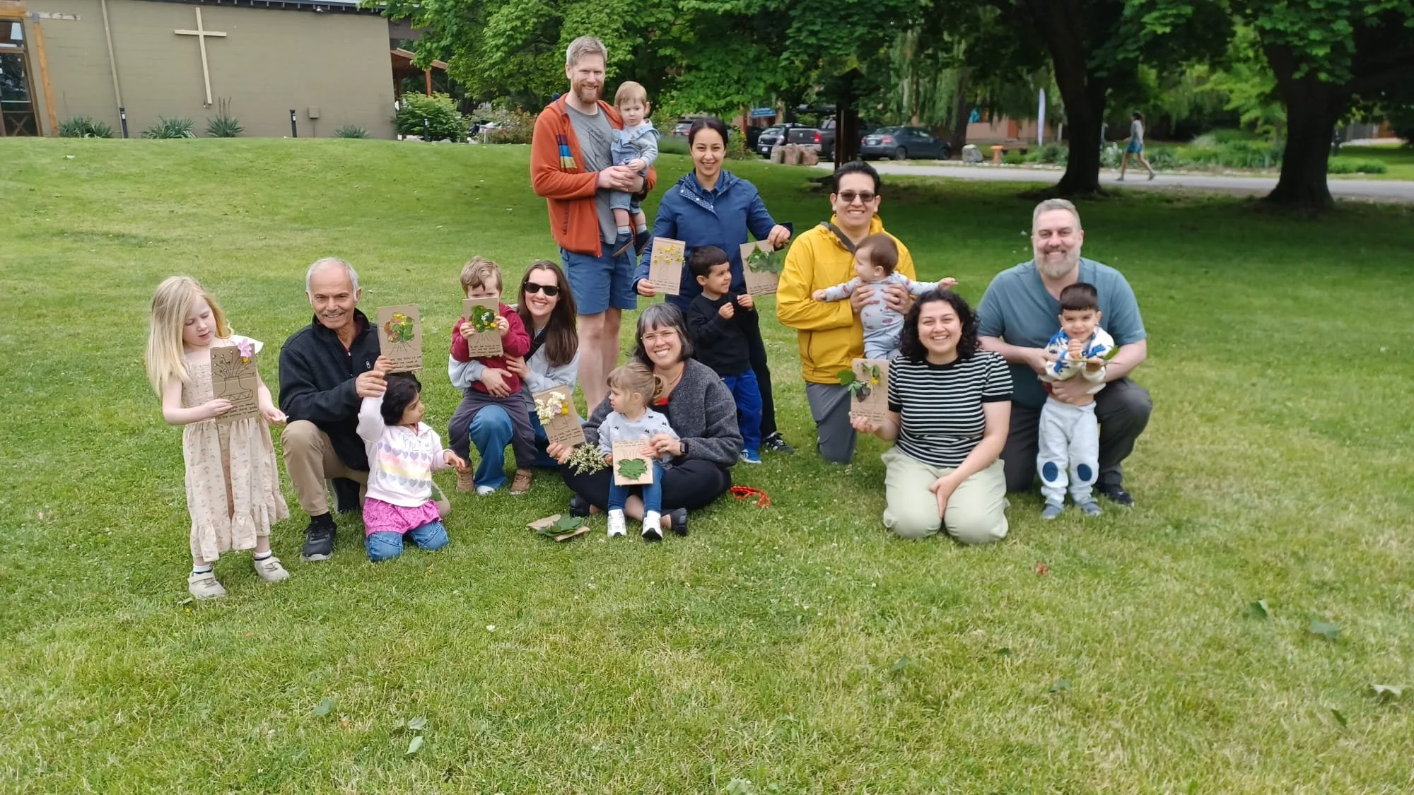 A group of people, including children and adults, gathered outdoors on a grassy area, holding hand-made cards or artwork with nature-themed designs. The group is smiling and posing for a photo, with trees and a building with a white cross in the background.