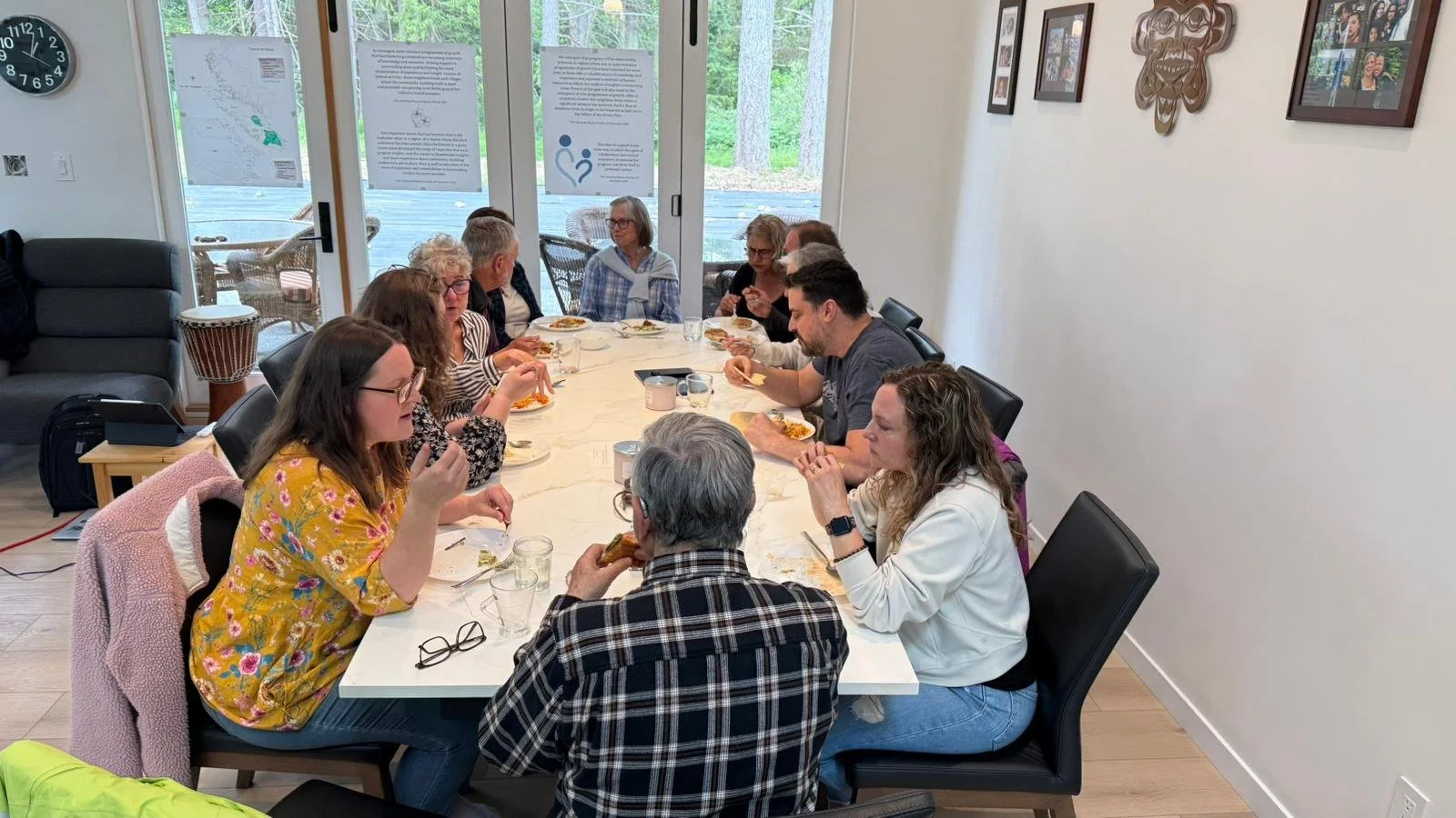 A group of people sitting around a rectangular dining table in a bright room, enjoying a meal together. The room has glass sliding doors with views of trees outside and framed photos on the wall.