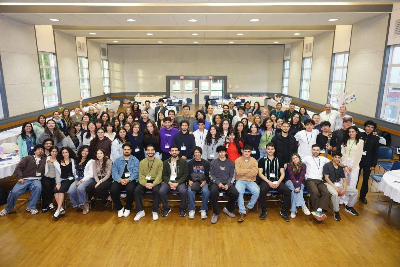 A large group of diverse young adults and some older adults gathered in a spacious conference hall for a group photo, with tables and windows visible in the background.
