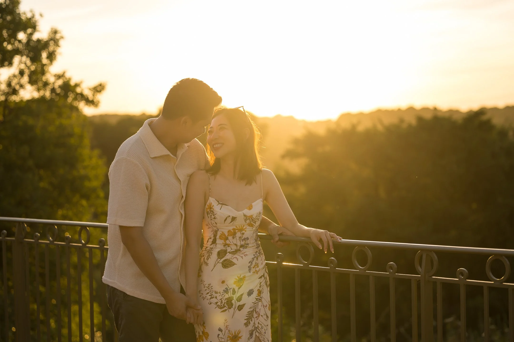 A couple is sharing a romantic moment outdoors at sunset, holding hands and leaning close to each other with a scenic background of trees and a sunset sky.