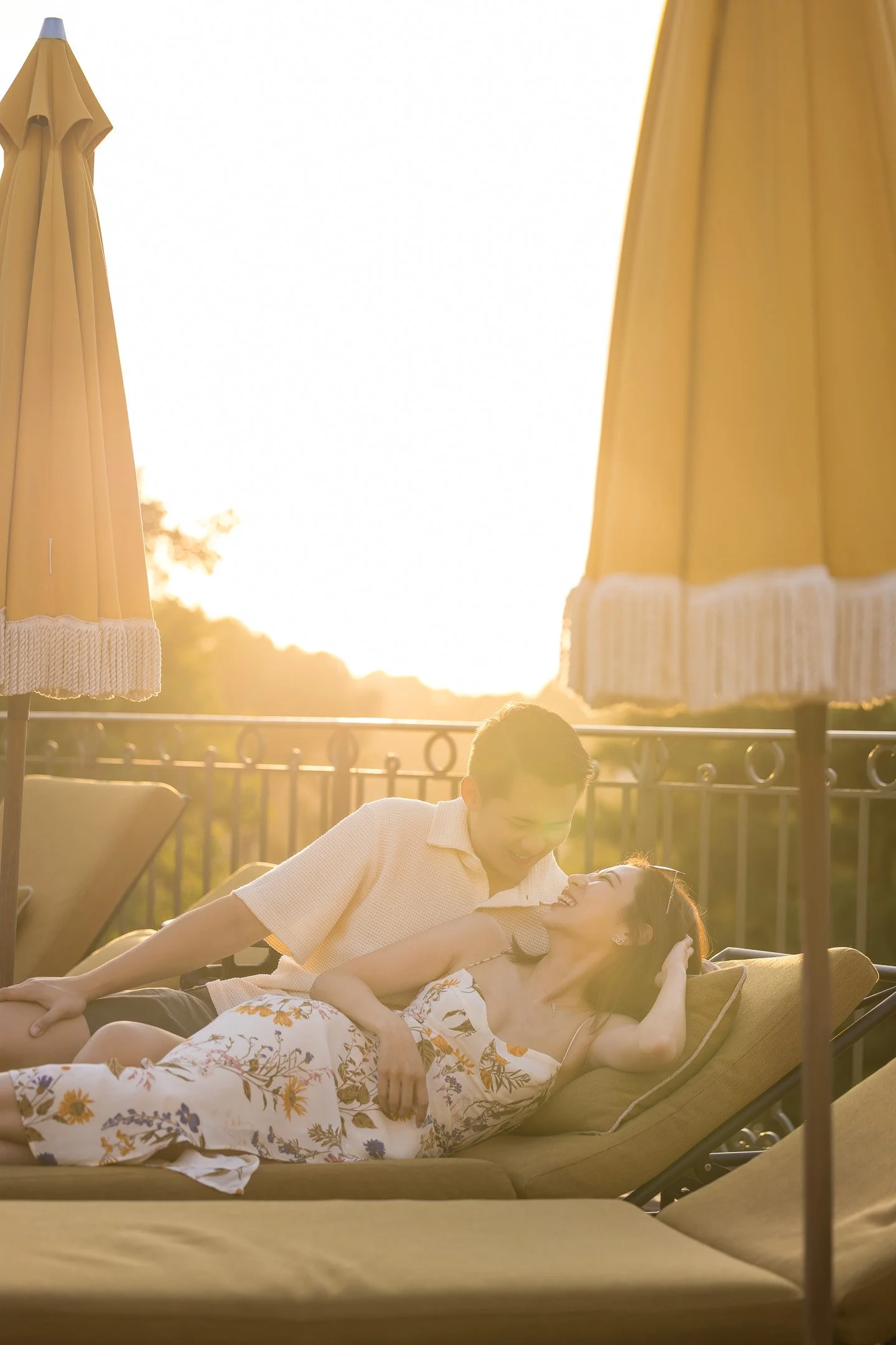 A couple is lying on a daybed outdoors during sunset, smiling and looking at each other, with yellow umbrellas in the background.