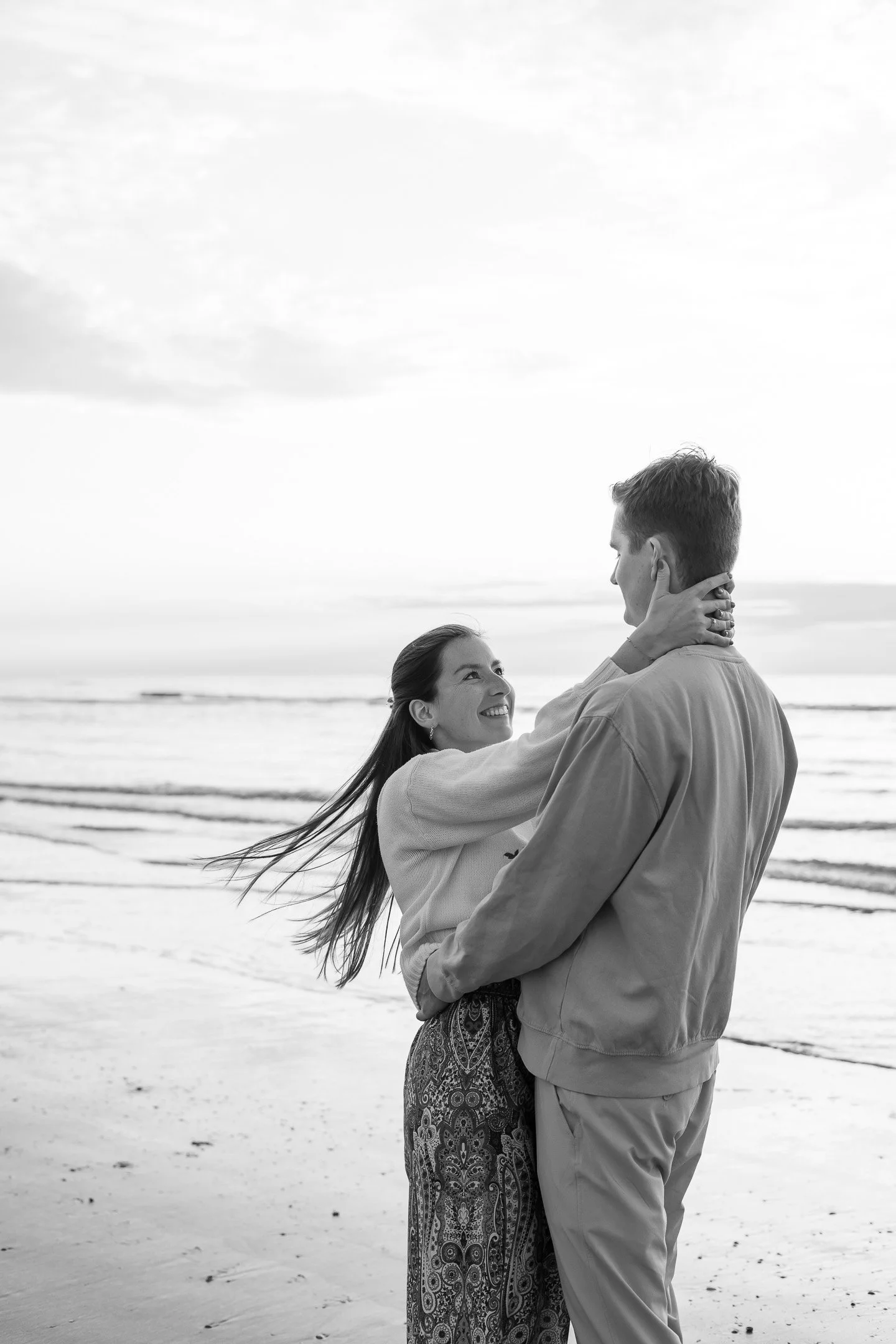 A couple stands on the beach with the ocean behind them, smiling and looking into each other's eyes; the woman is holding the man's face while they embrace.