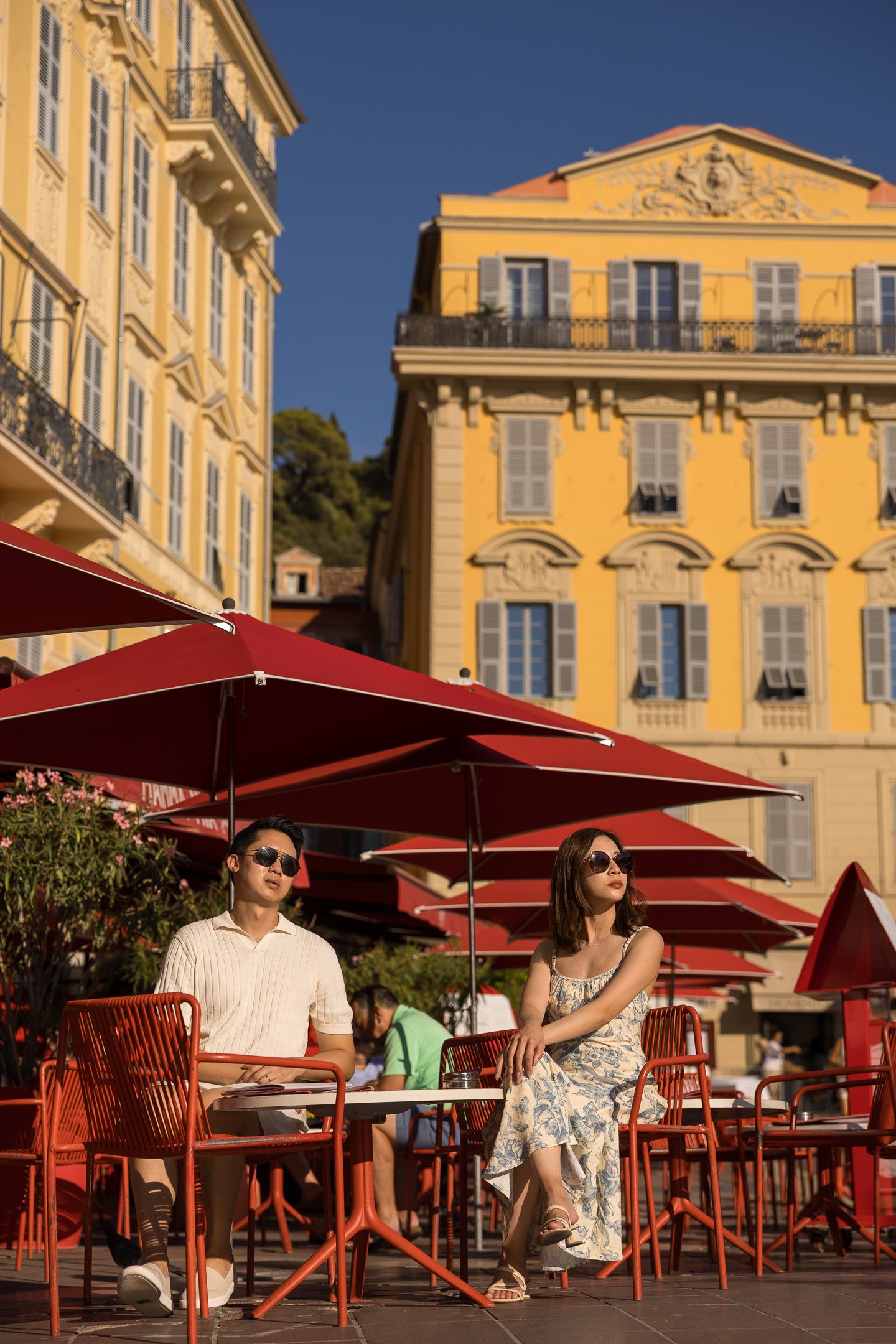Two people, a man and a woman, sitting at an outdoor cafe with red umbrellas on a sunny day. The buildings behind them are painted yellow with multiple windows, some open, and classic European architecture.