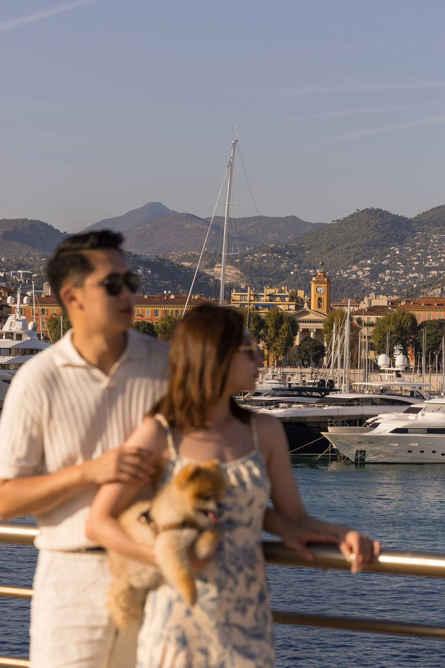 People walking along a marina with boats, mountains in the background, and a clock tower in a coastal city.