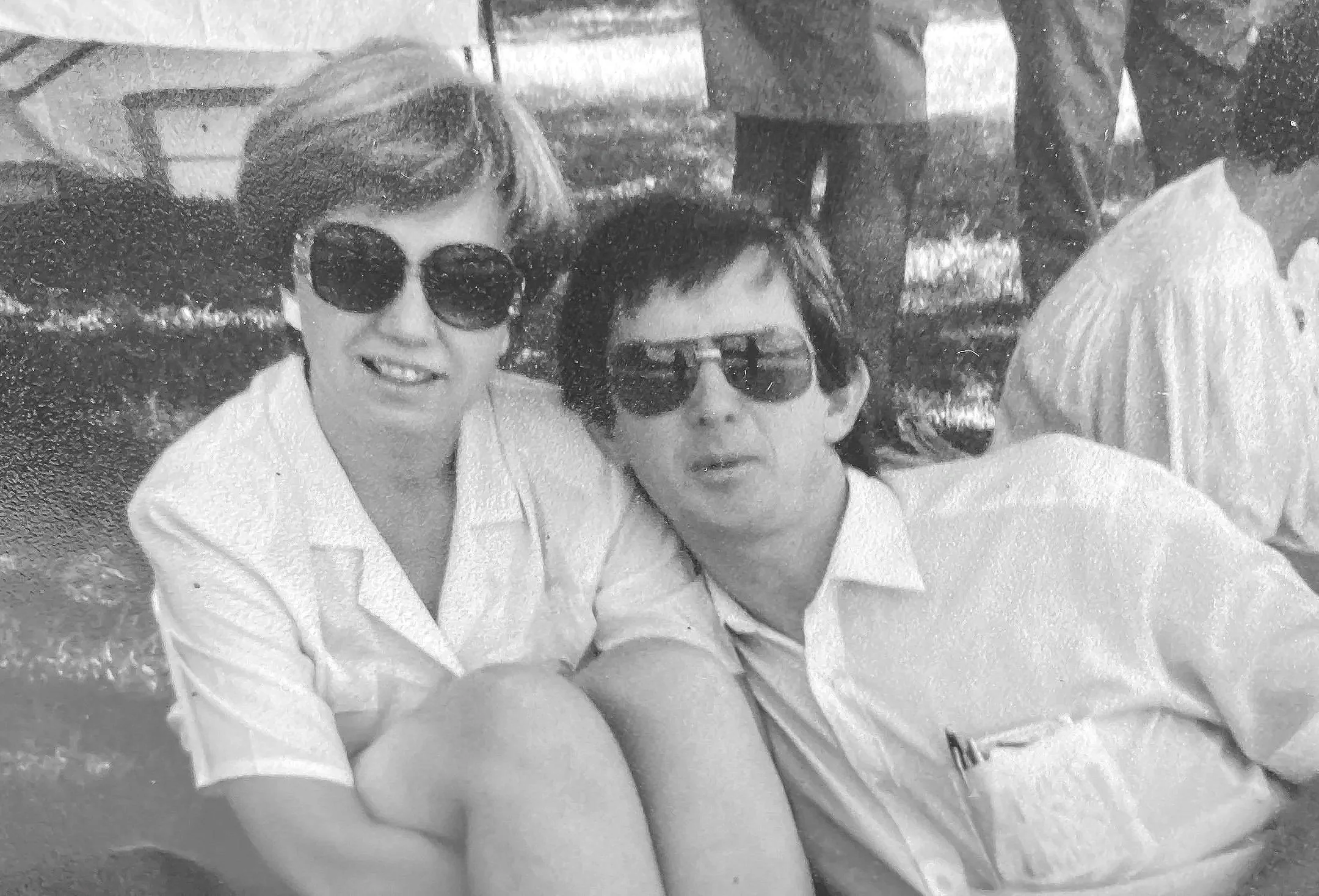 Two women sitting outdoors, both wearing white shirts and sunglasses, smiling for the photo. One woman has short hair and the other has medium-length hair. The background includes blurred trees and a parked vehicle.