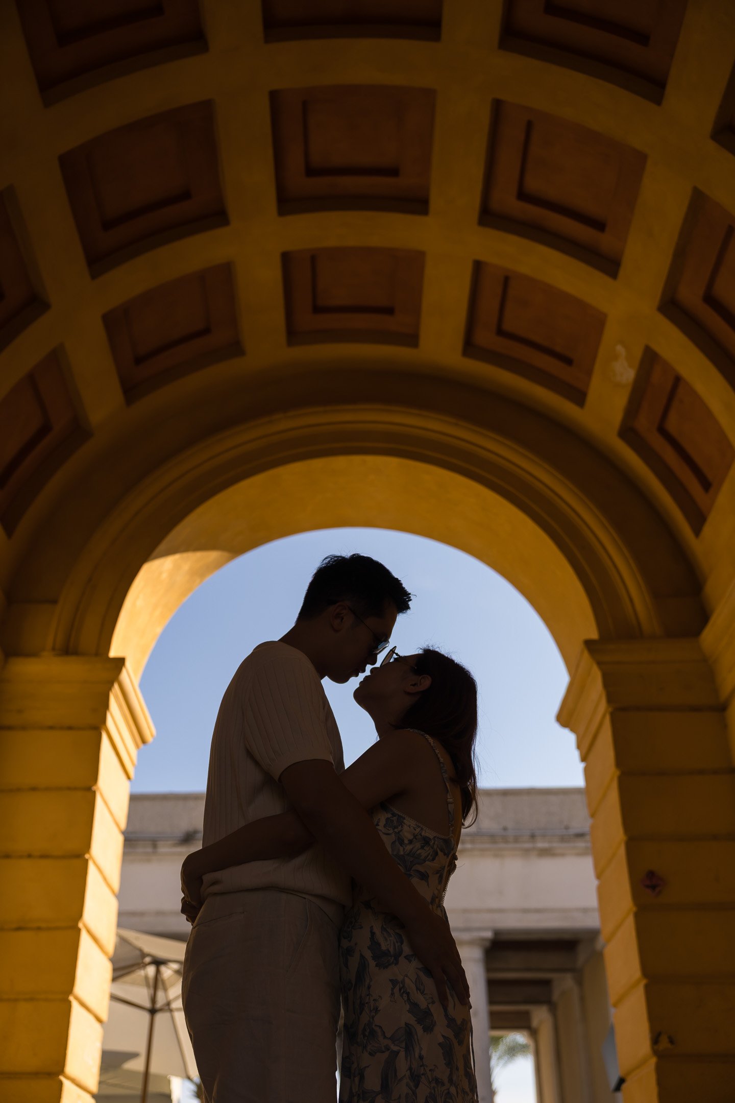 A couple standing close together under a yellow archway, with the sky visible behind them.