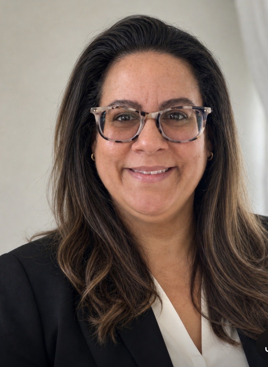 A woman with shoulder-length brown hair wearing glasses, a black blazer, and a white blouse, smiling in front of a plain background.