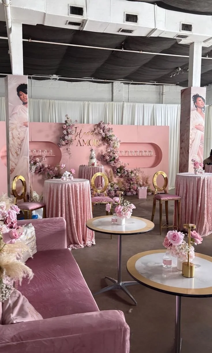 Decorated event space with pink and gold accents, featuring floral arrangements, round tables with pink tablecloths, and a backdrop with the words 'Island Amour' and a cake in the center.