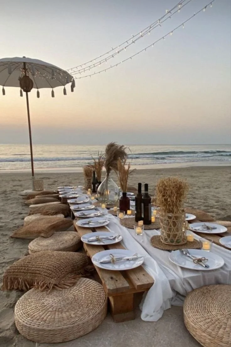Beachside dining setup with a long table, white plates, cutlery, candles, and decorative vases, surrounded by woven cushions and pillows under a large umbrella, with the ocean and a sunset in the background.