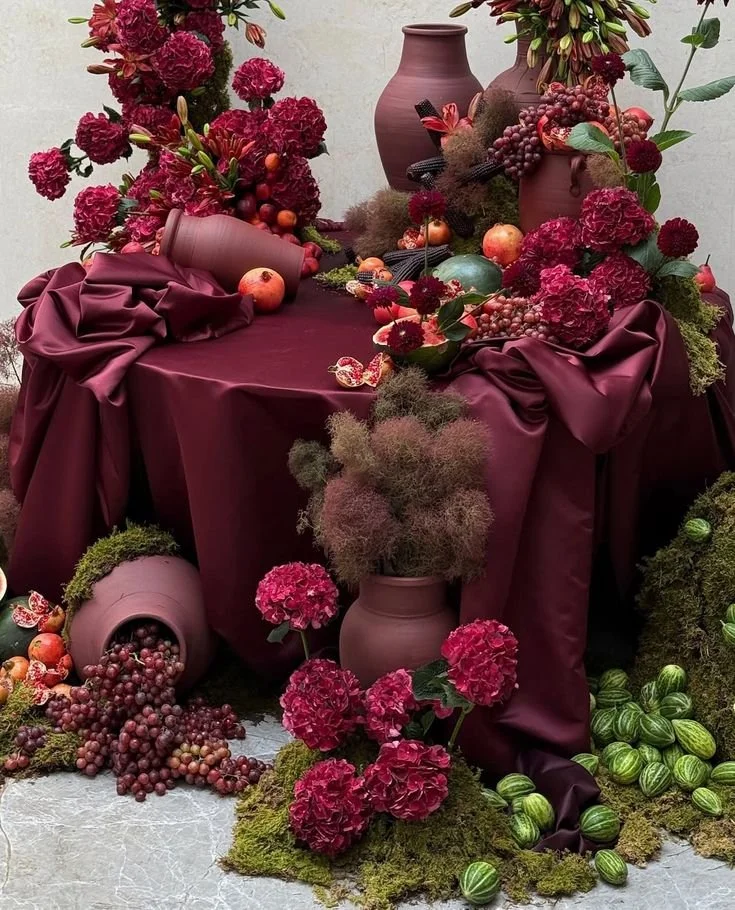 A decorative table display with vases, flowers, and fruits, including grapes, apples, and watermelons, with a burgundy satin tablecloth and floral arrangements mainly in red, pink, and green.